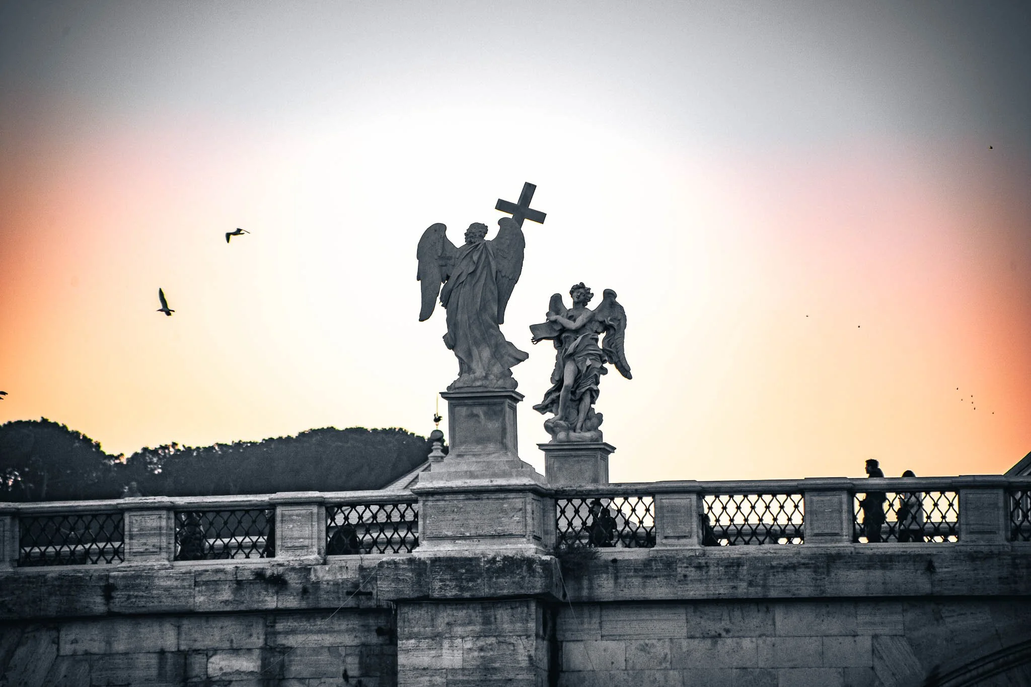 Statues of angels on a bridge at sunset, with birds flying in the sky and people walking in the background.