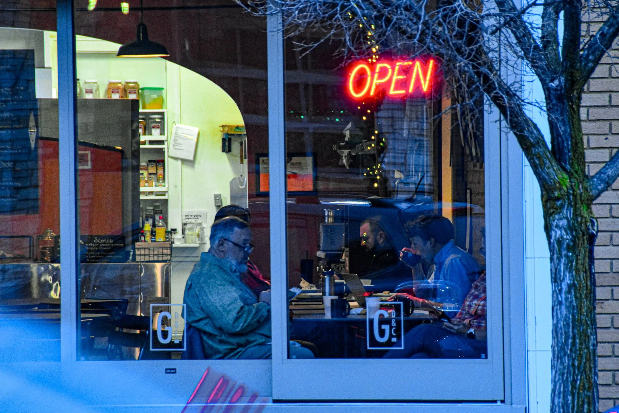 People sitting inside a cafe, visible through a large glass window, with an illuminated red "Open" sign and a tree outside. Inside, three individuals are working on laptops, with various supplies and a microwave in the background.