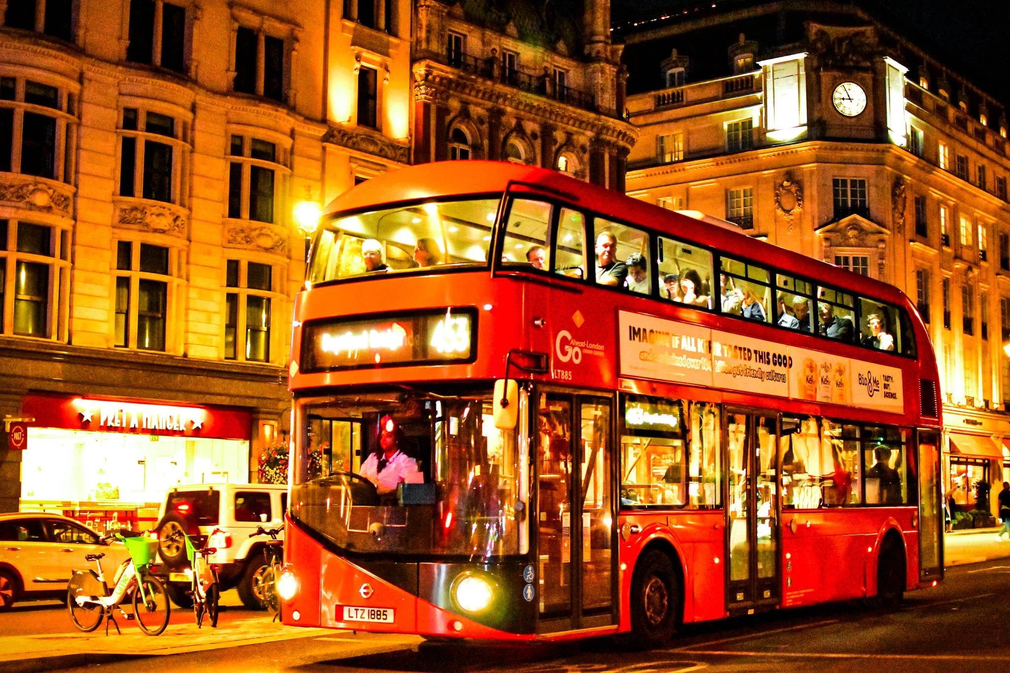 A red double-decker bus with passengers on the upper deck travels through a city street at night, illuminated by streetlights and building lights, with cars and bicycles nearby.