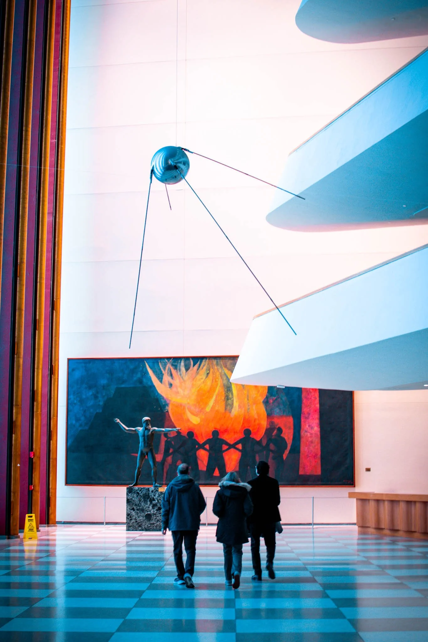 Four visitors walk through an art gallery with a large abstract painting of a fire and silhouette figures in the background. The gallery features modern architecture, including white angular structures and a suspended spherical sculpture.