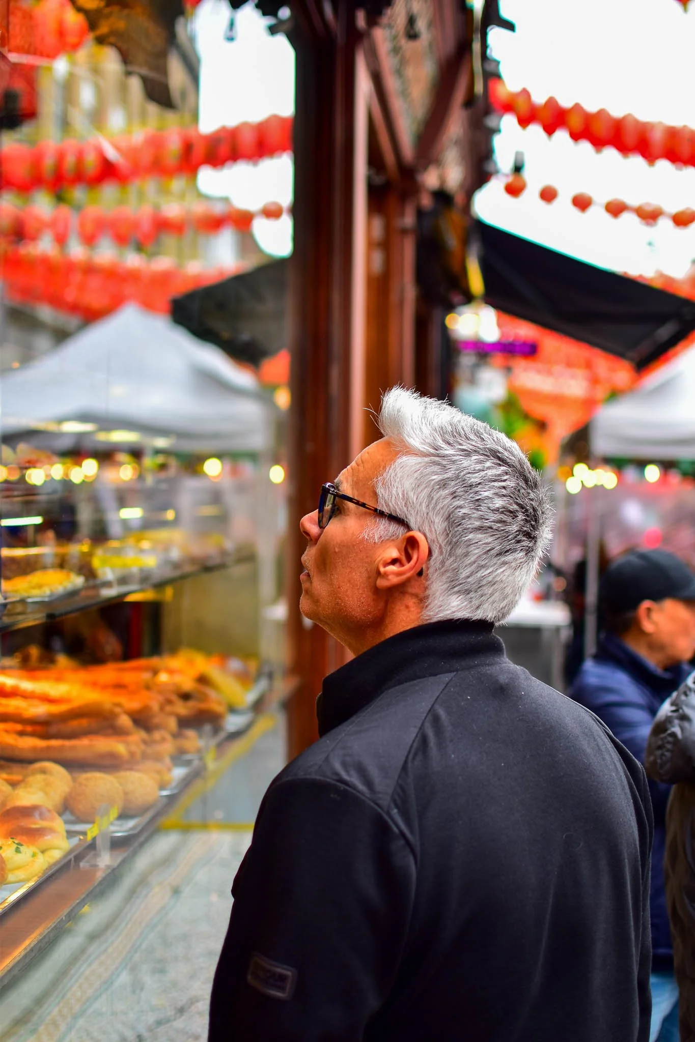 An older man with gray hair and black glasses looks at a display of baked goods at an outdoor market decorated with red lanterns.