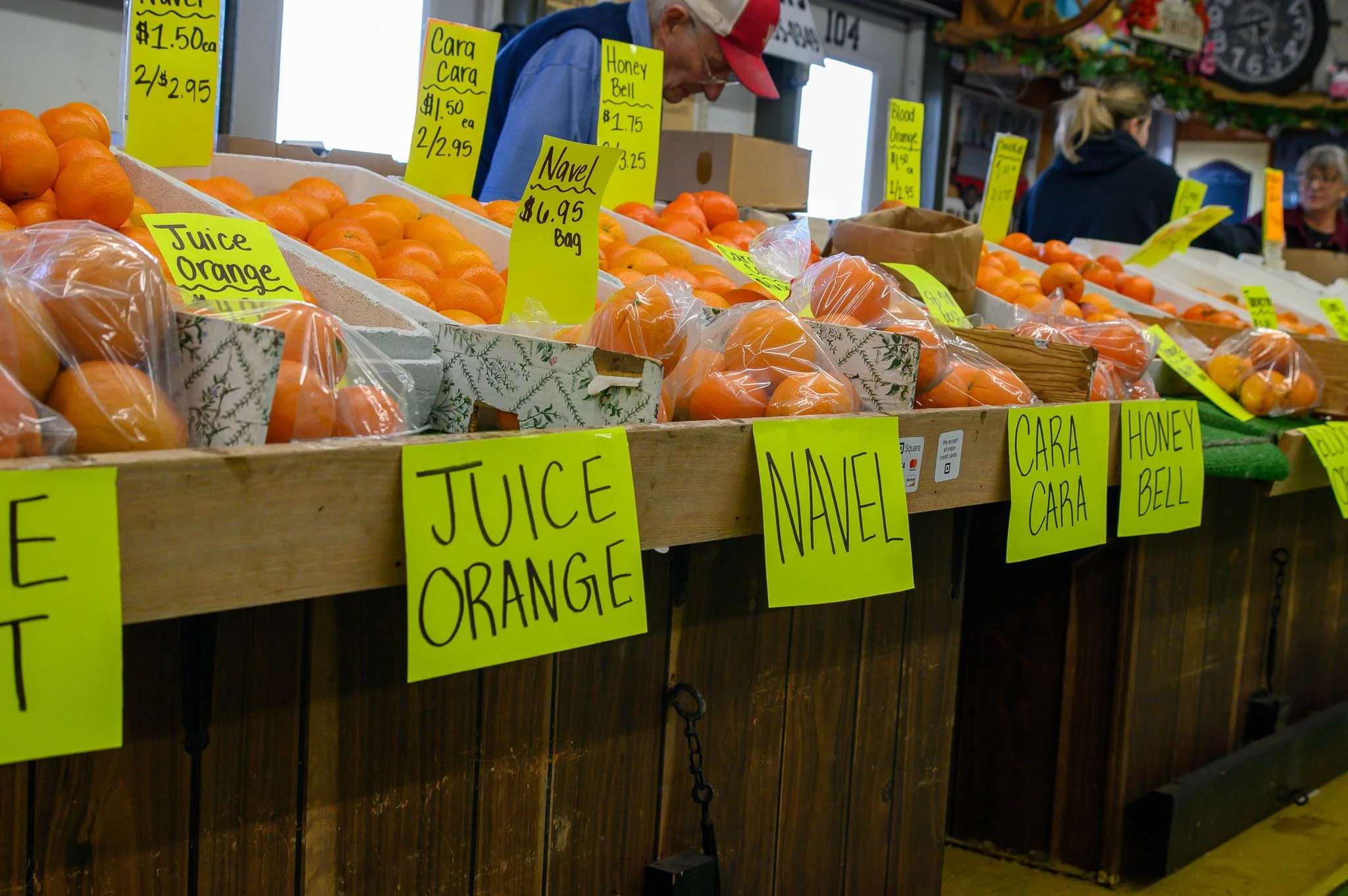 A display of oranges at a market stall with yellow handwritten signs indicating prices and types, including juice oranges, navel oranges, honey bell, and cara cara oranges, with several people shopping in the background.