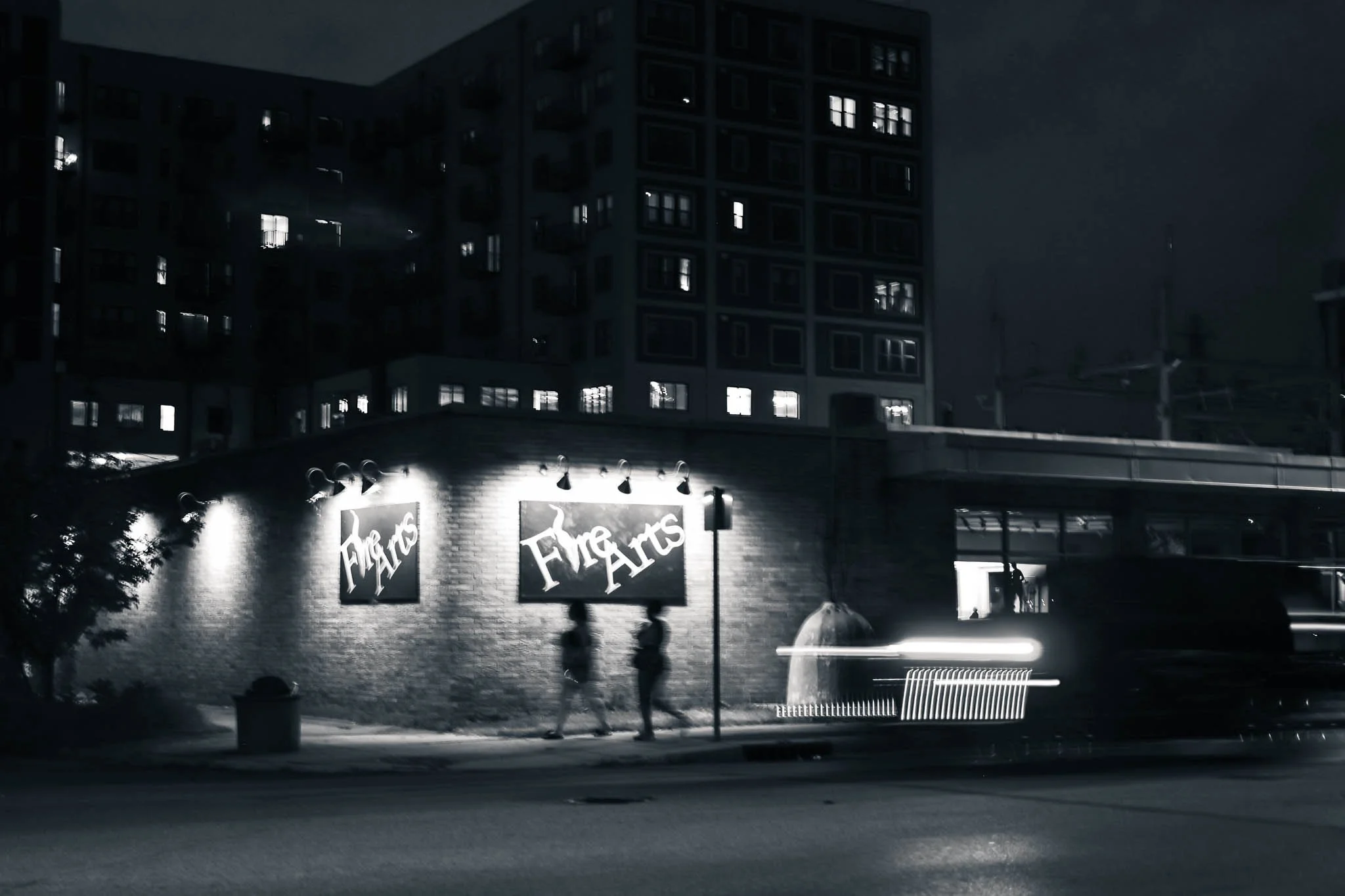 Nighttime scene of a brick building with two illuminated signs reading 'Fire Arts' and people walking along the sidewalk. A bench, a trash can, and a parked car with light streaks from passing vehicle headlights are visible in the foreground.