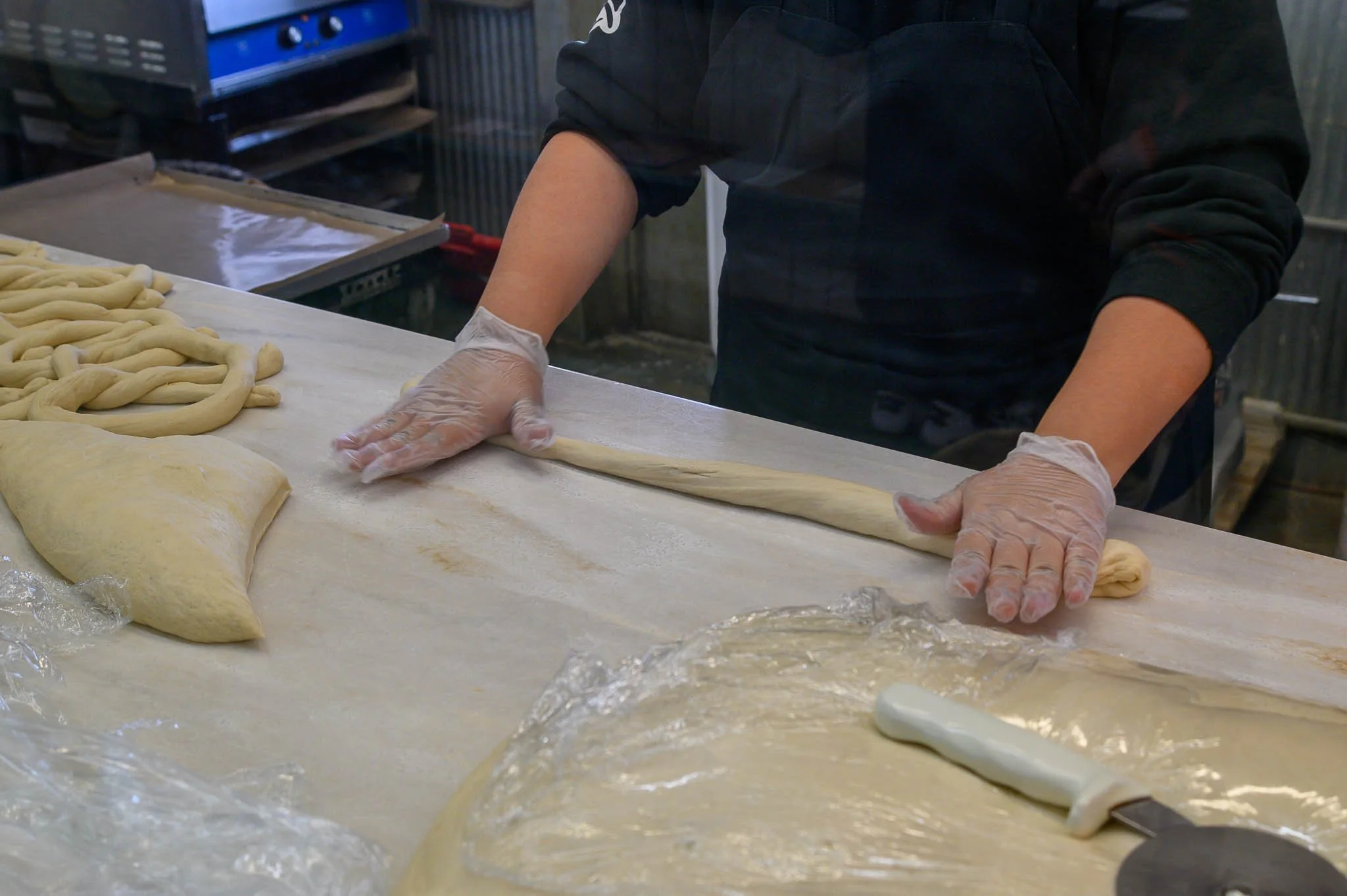 Person in black clothing and gloves rolling out dough on a bakery counter with more dough and a knife nearby.