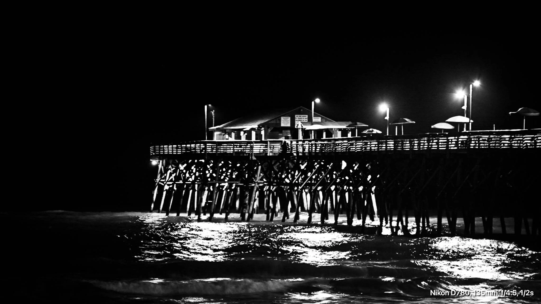 Night view of a pier with lights, supporting structures, and a building at the end, extending into the dark ocean, with waves reflecting light.