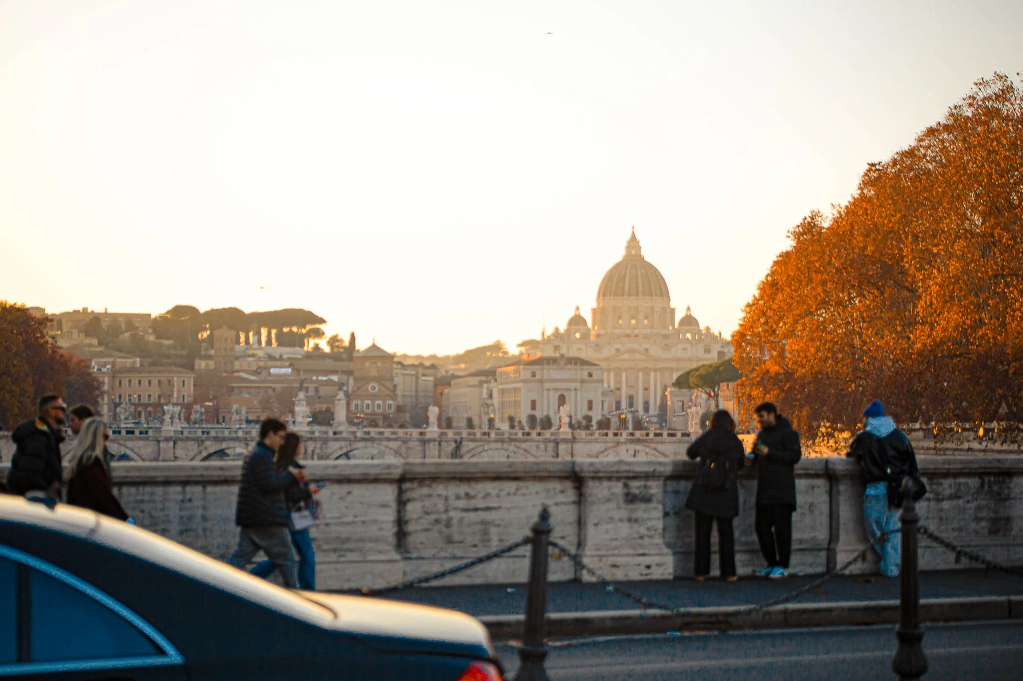 People standing and walking along a bridge with a view of St. Peter's Basilica in Vatican City, during sunset with orange trees on the right.