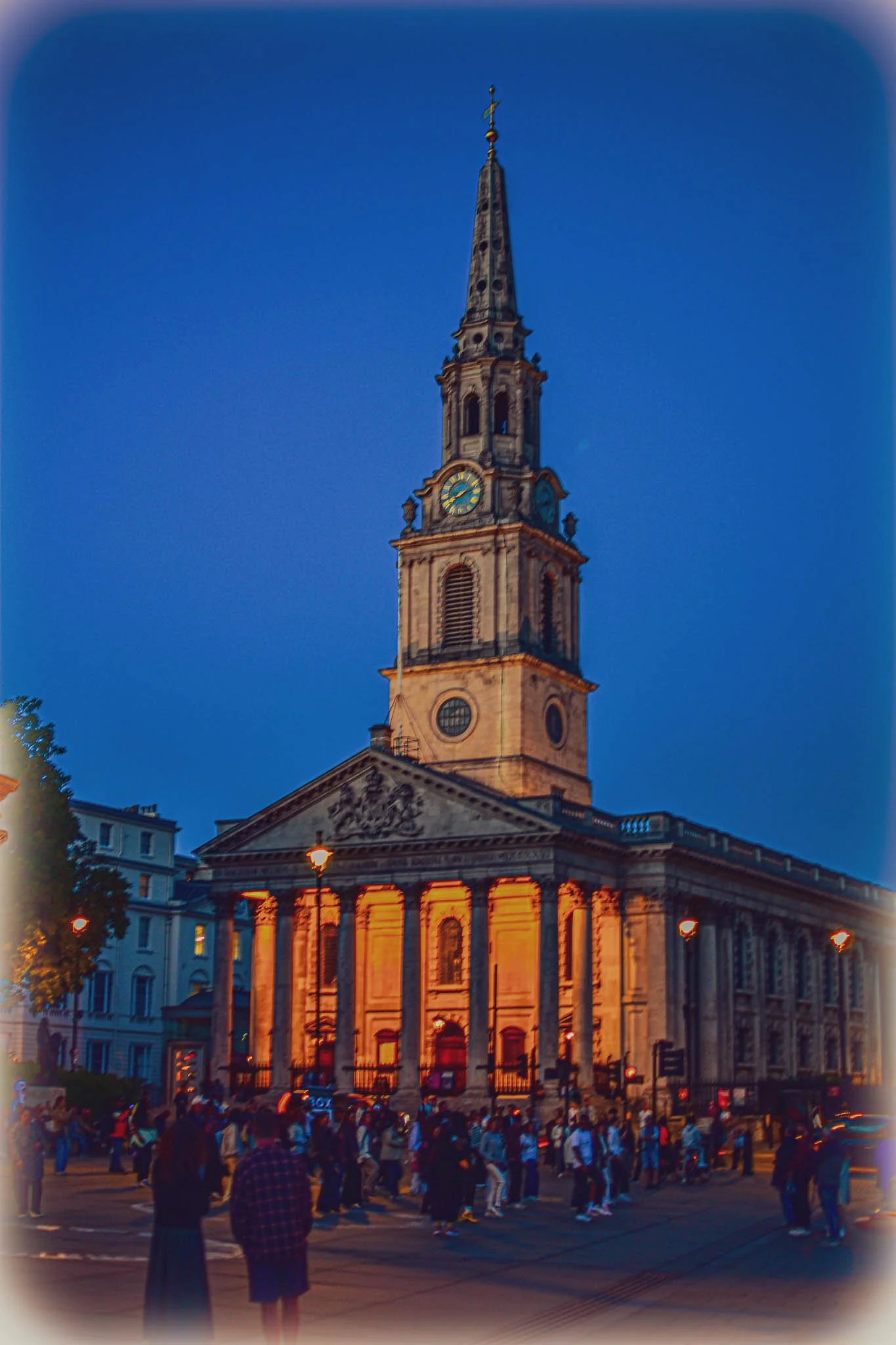 Historical church with a tall spire illuminated at dusk, with a crowd of people in front.