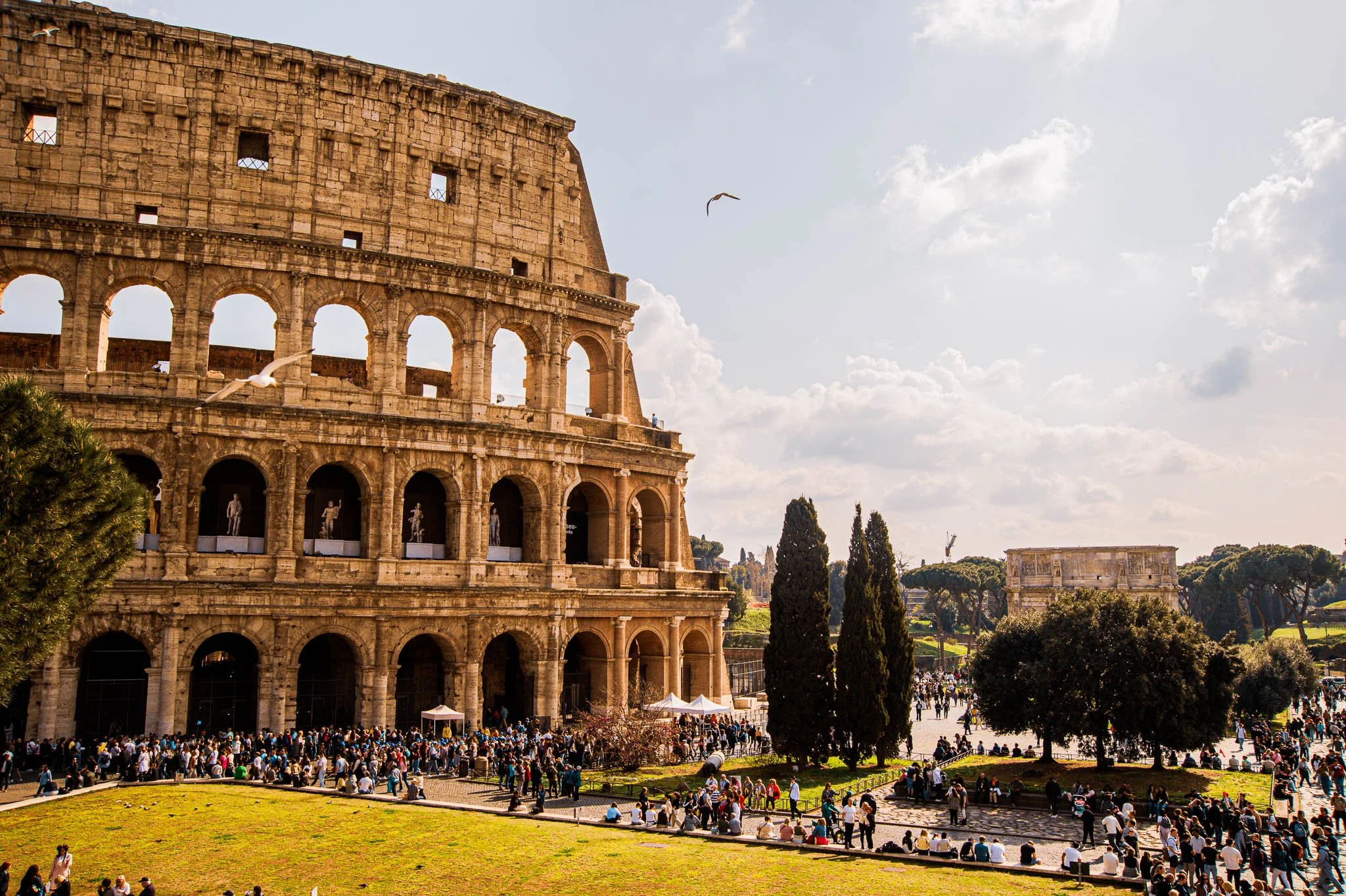 The Colosseum in Rome, Italy, with visitors gathering outside and some walking around, facing a partly cloudy sky with seagulls flying overhead.
