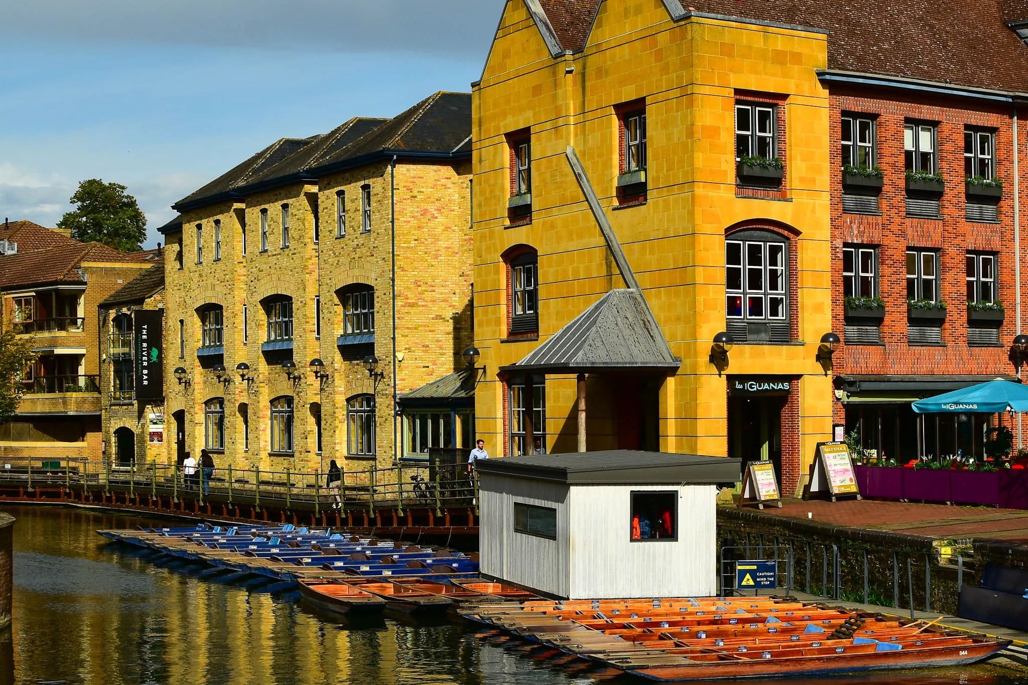 Colorful buildings along a waterway with small boats docked in the foreground, and a wooden walkway with people walking.