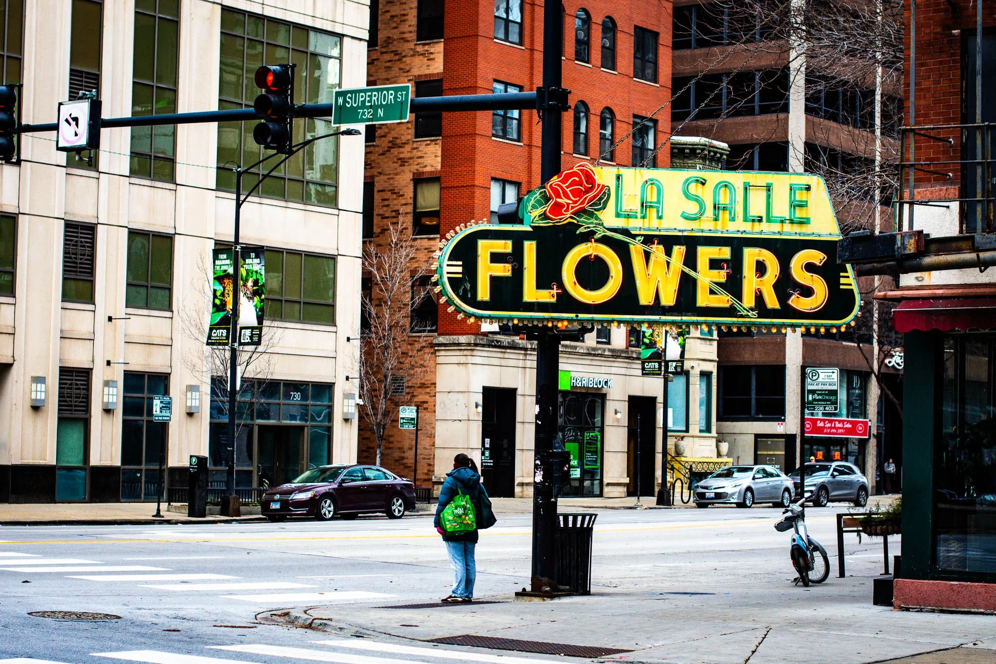 A street scene showing a sign for La Salle Flowers, a woman standing at the corner with a green backpack, cars on the street, and buildings in the background.
