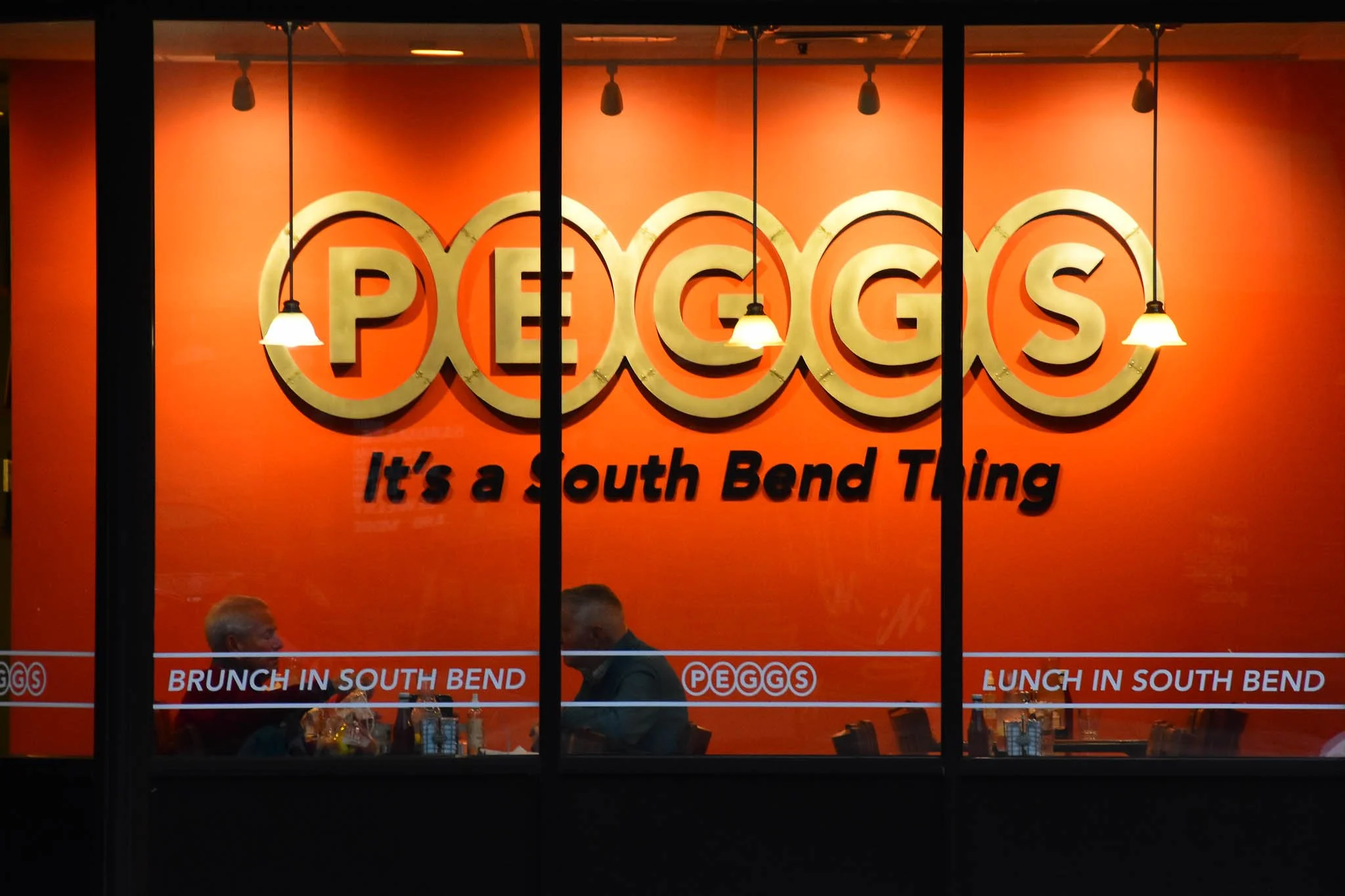Restaurant window display for Peggs in South Bend with the slogan "It's a South Bend Thing," showing two people dining inside and text advertising brunch and lunch services.