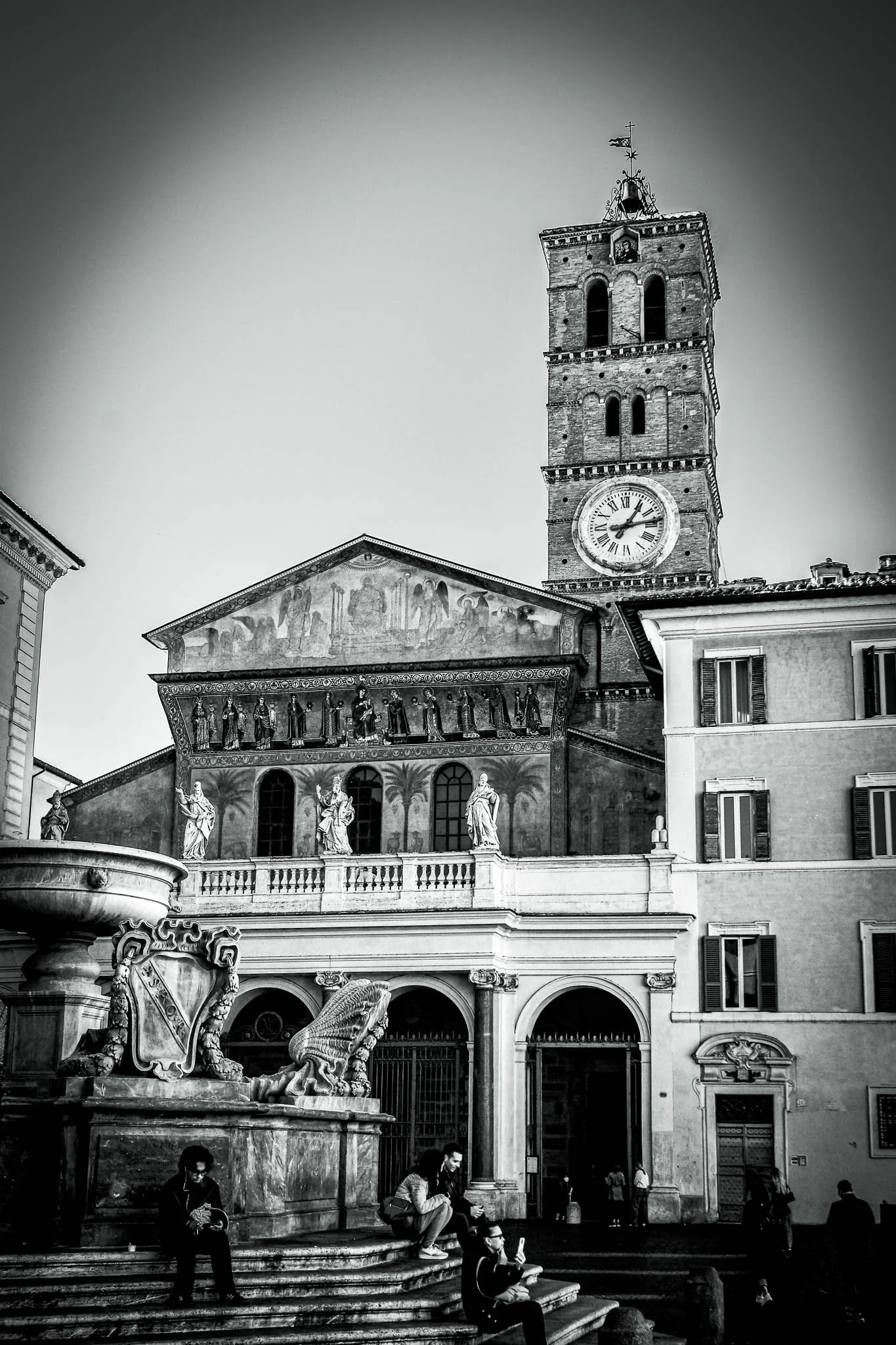 Black and white photo of a historic clock tower and church with statues and people sitting on steps in a European city.