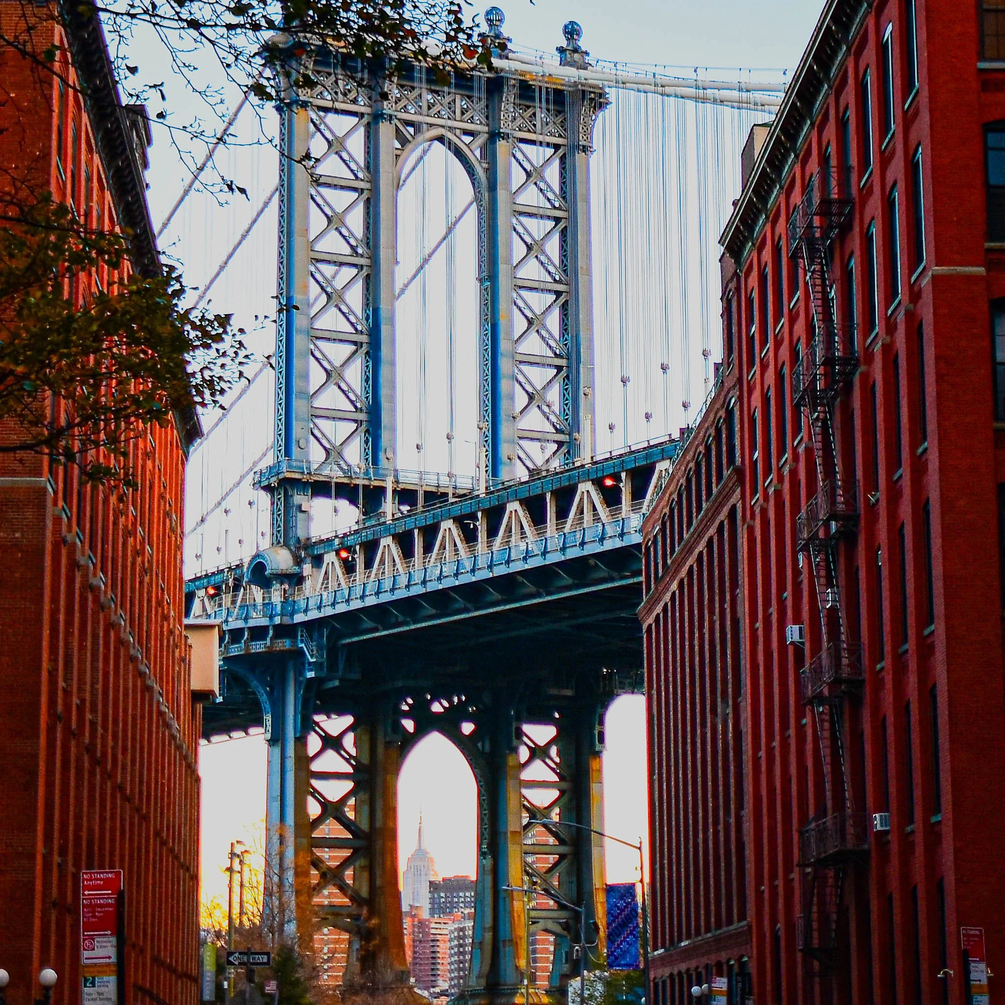 The image depicts the Brooklyn Bridge in New York City, viewed from a street level between red-brick buildings, with the Empire State Building visible through the bridge's arch in the background.