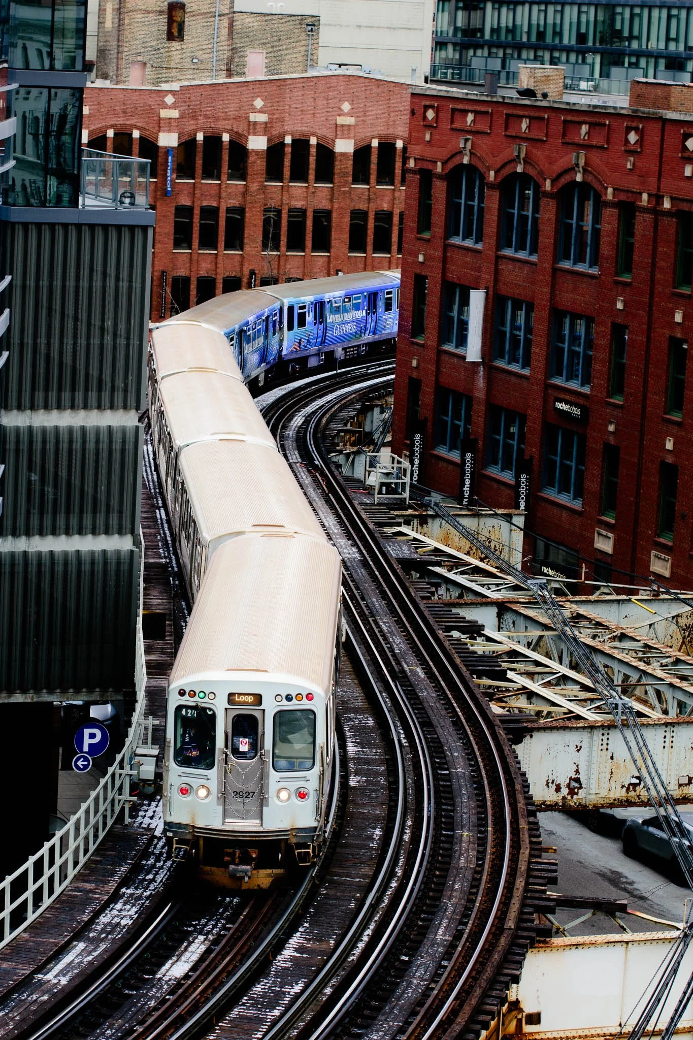 An elevated view of a train on a curved track passing between tall brick buildings in an urban setting.