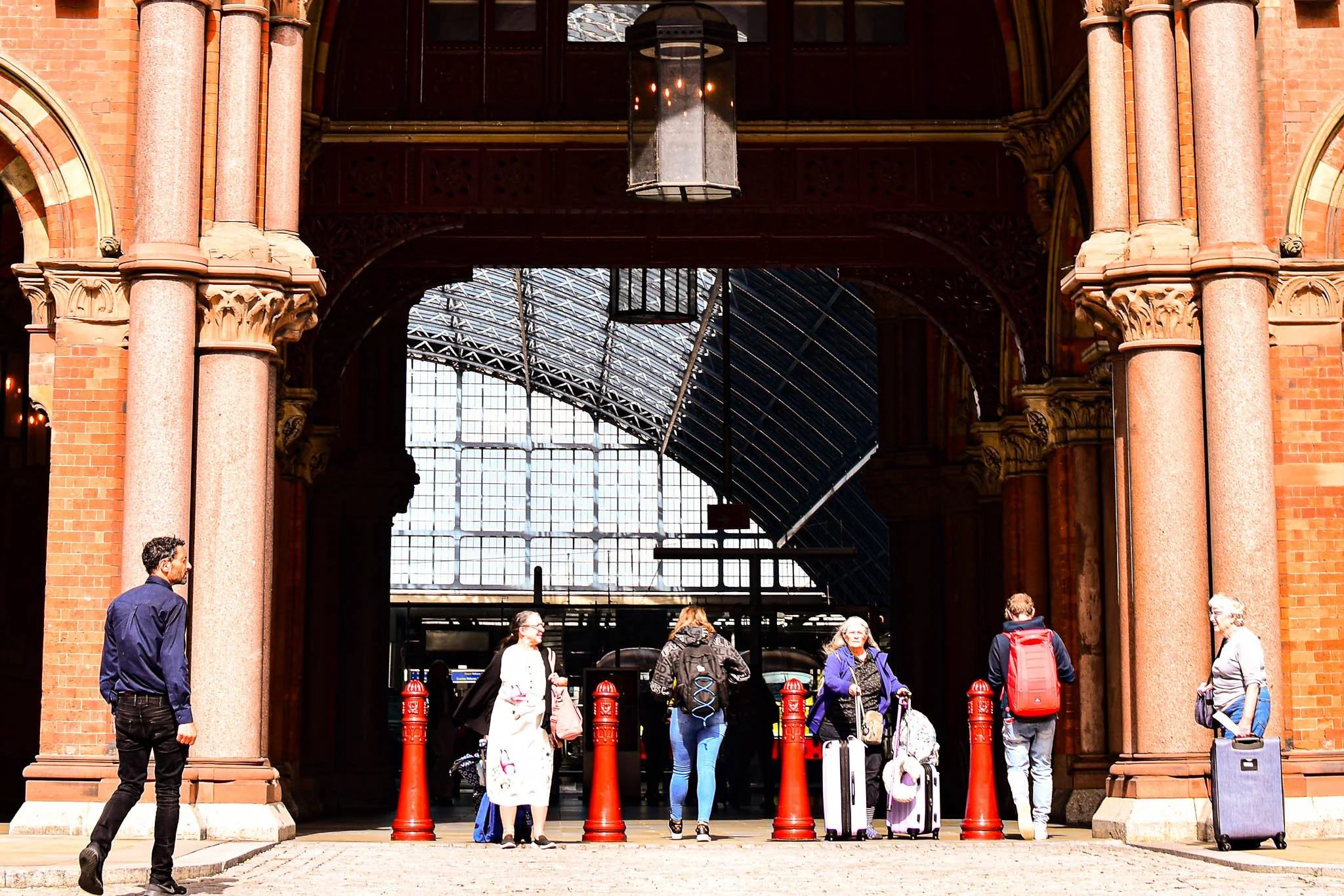 People with luggage waiting at a train station entrance framed by red brick columns and an arched glass roof.
