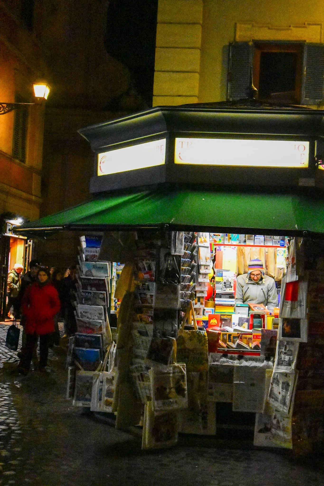 Night scene of a street bookstore kiosk selling newspapers, magazines, and books, illuminated under a green canopy, with pedestrians walking by.