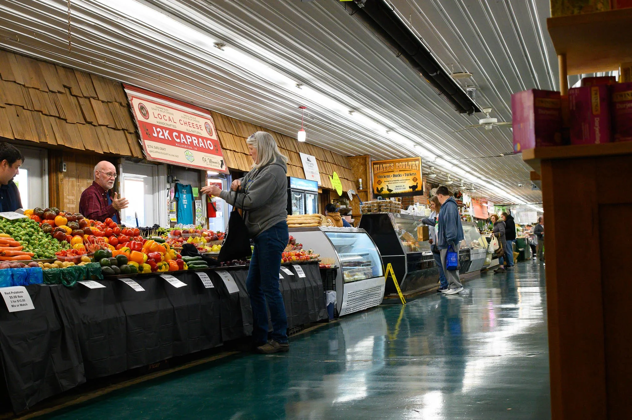People shopping for fresh produce at a market with fruit and vegetable stalls, and signs for local cheese and poultry in the background.