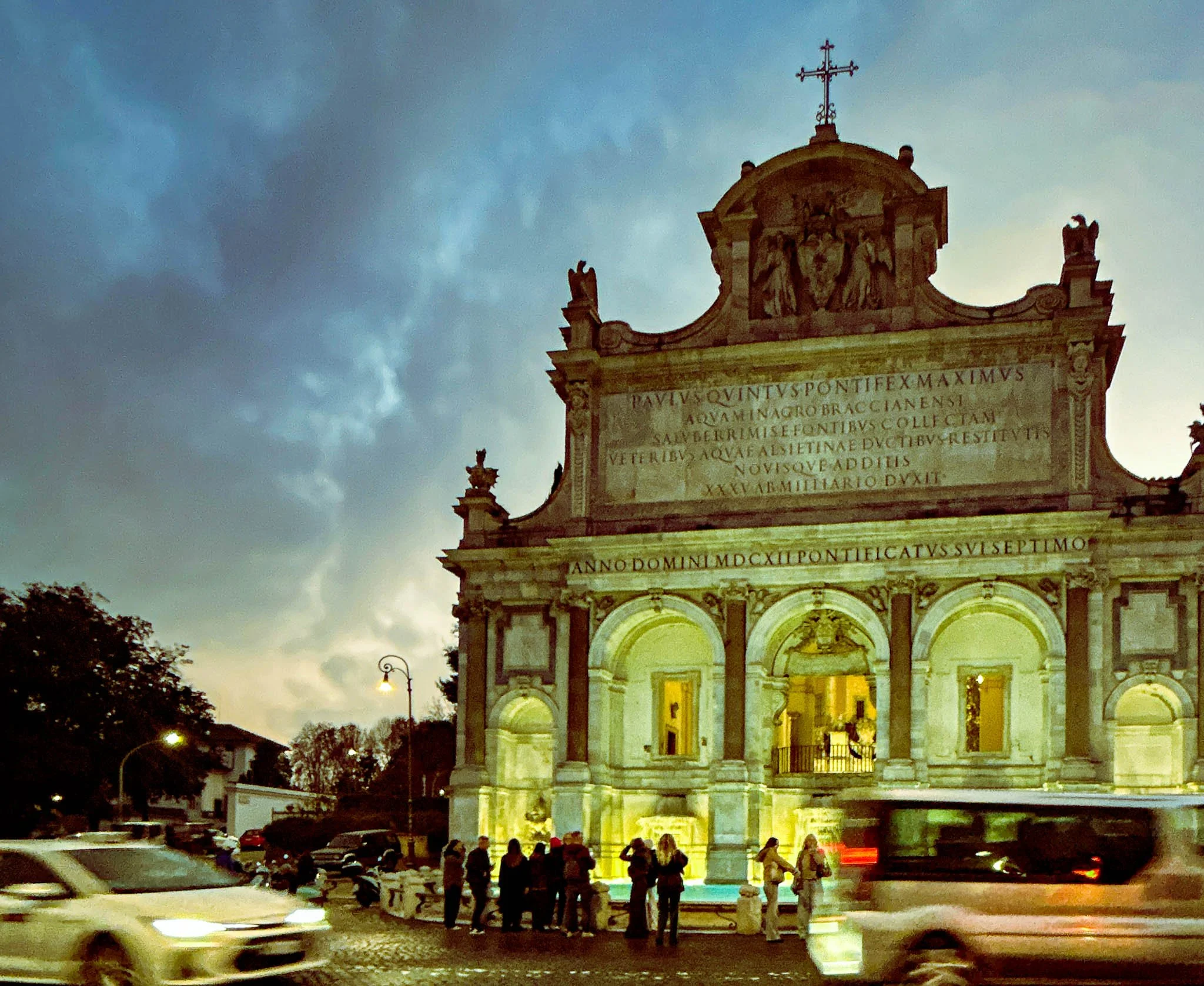 Baroque church facade illuminated at dusk with people gathered in front, cars passing by, and cloudy sky overhead.