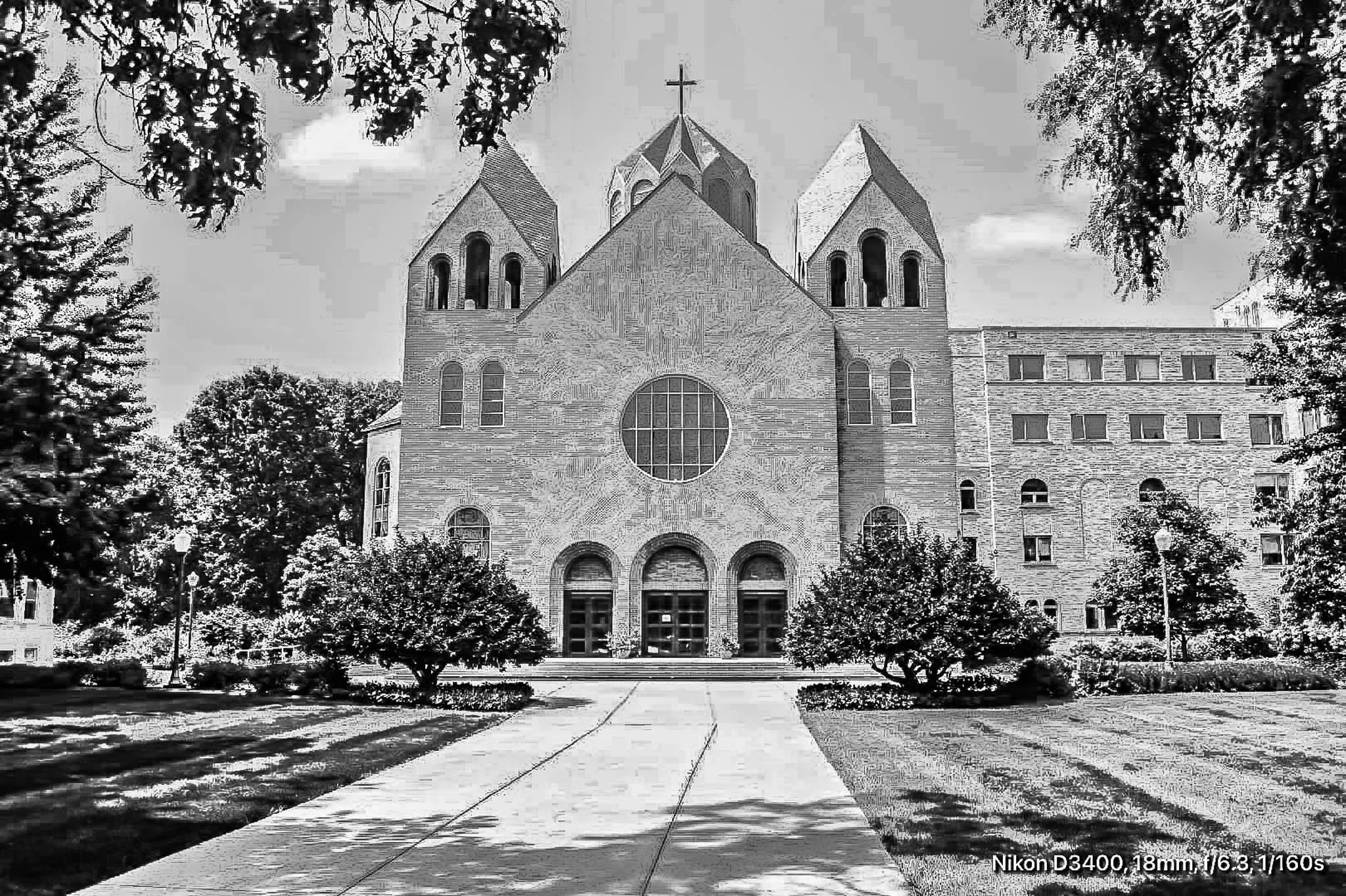 Black and white photo of a church with two towers, a large window, and three arched doors, surrounded by trees and a pathway leading to the entrance.