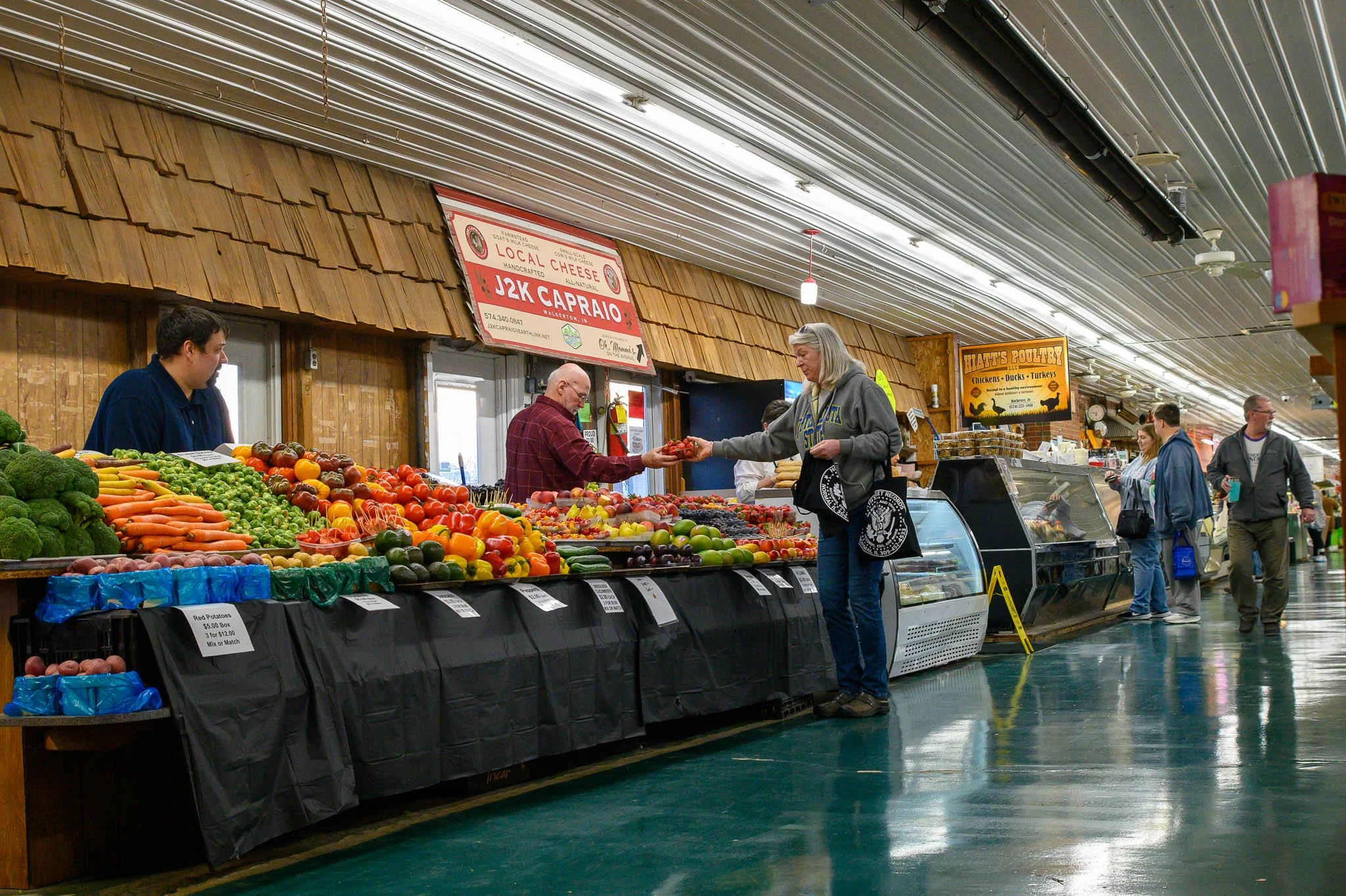 Inside a grocery store or farmers market, a woman is purchasing fresh vegetables from a vendor at a produce stand. There are various vegetables like broccoli, carrots, and tomatoes on display. Other shoppers are browsing in the background.