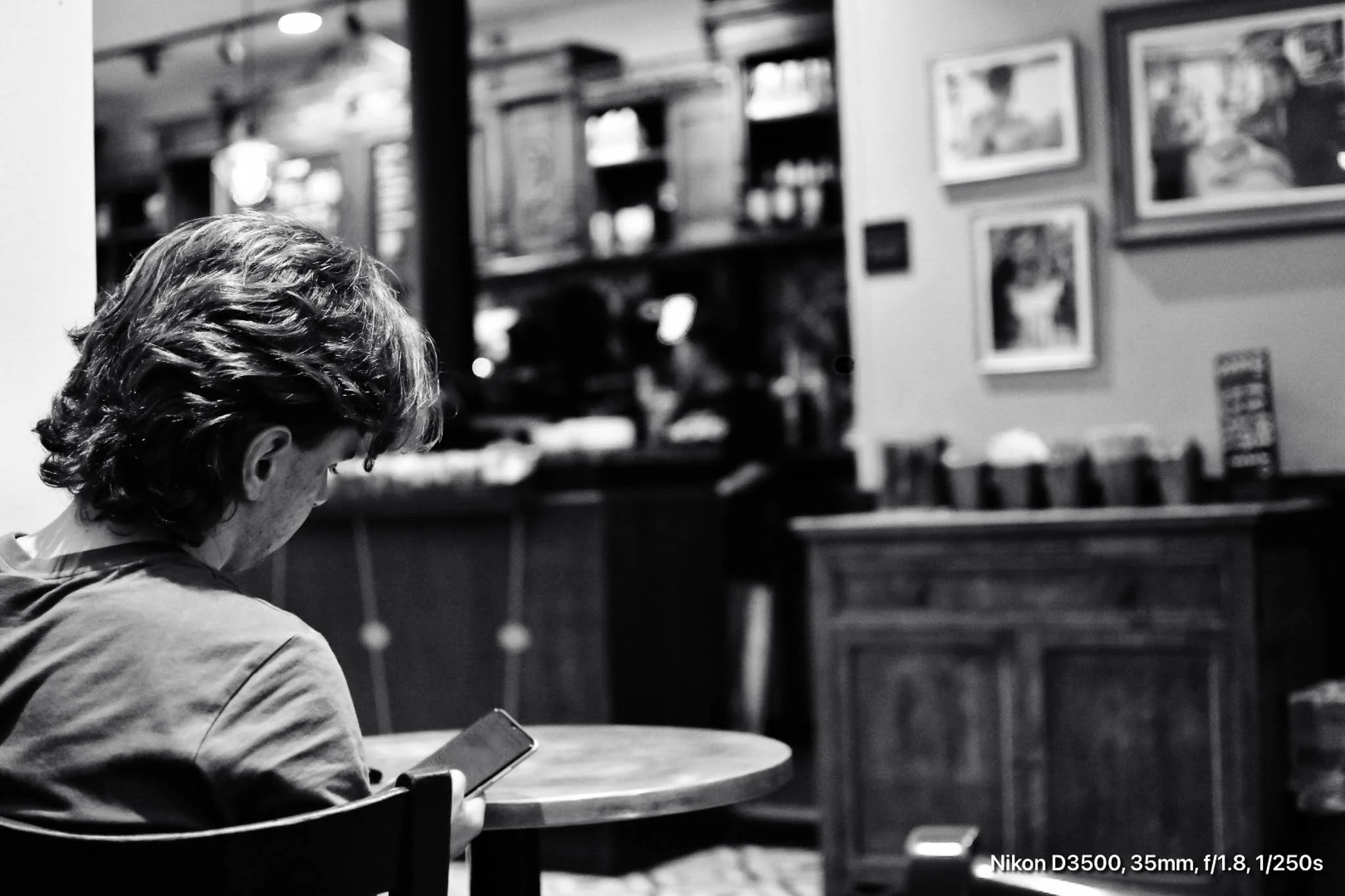 A person with wavy hair sitting at a round table using a smartphone in a cafe or restaurant with framed pictures on the wall in the background.