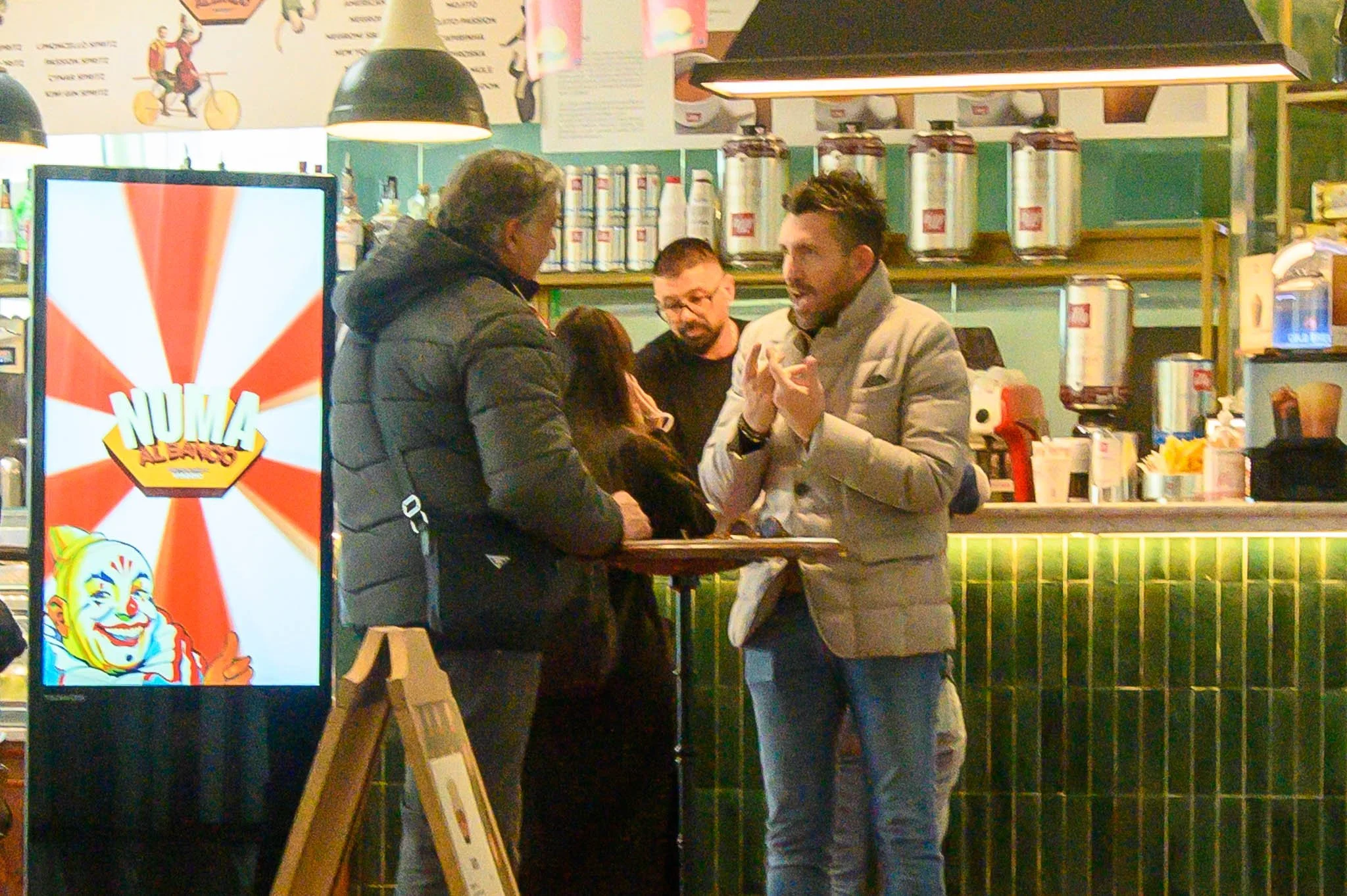 Four people, two men and two women, are standing at a counter inside a food establishment, engaging in a conversation. One man is wearing a light gray blazer and gesturing with his hands, while the others listen. There are various containers and equi