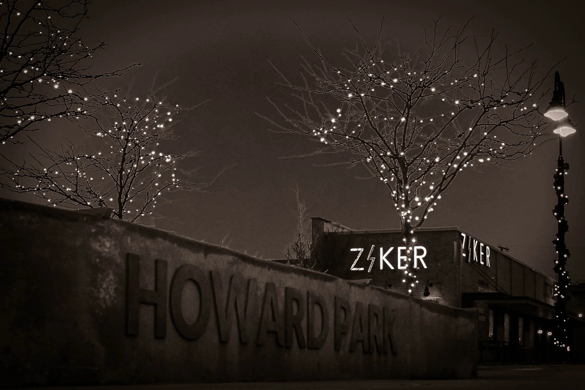 Night view of Howellard Park with trees decorated with string lights, a sign reading 'Z¥KER,' and ambient street lighting.