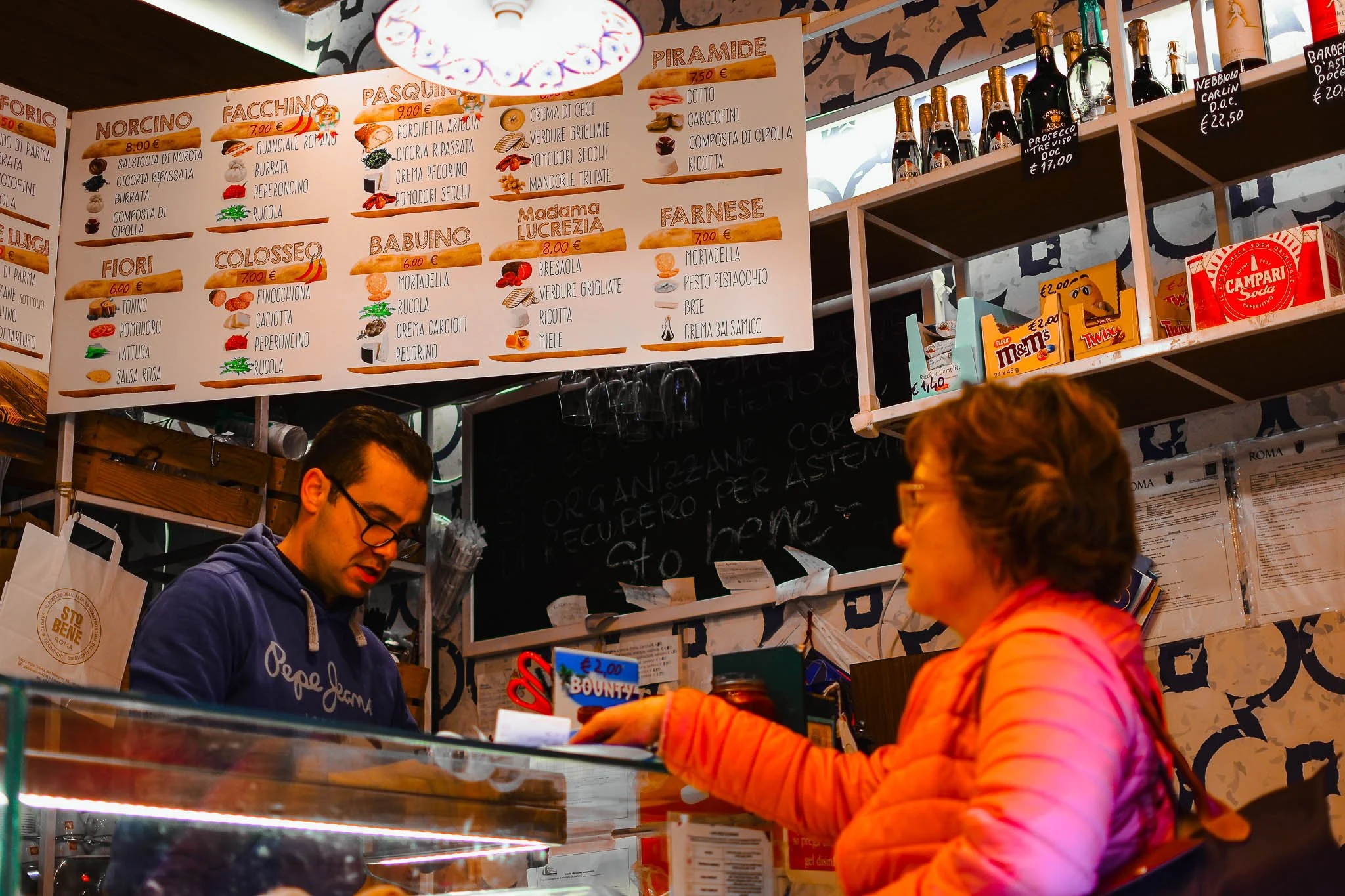 Customer ordering at a counter in an Italian deli or cafe with a large menu board overhead displaying various food and drink options, and shelves of candies and bottles of drinks behind the counter.