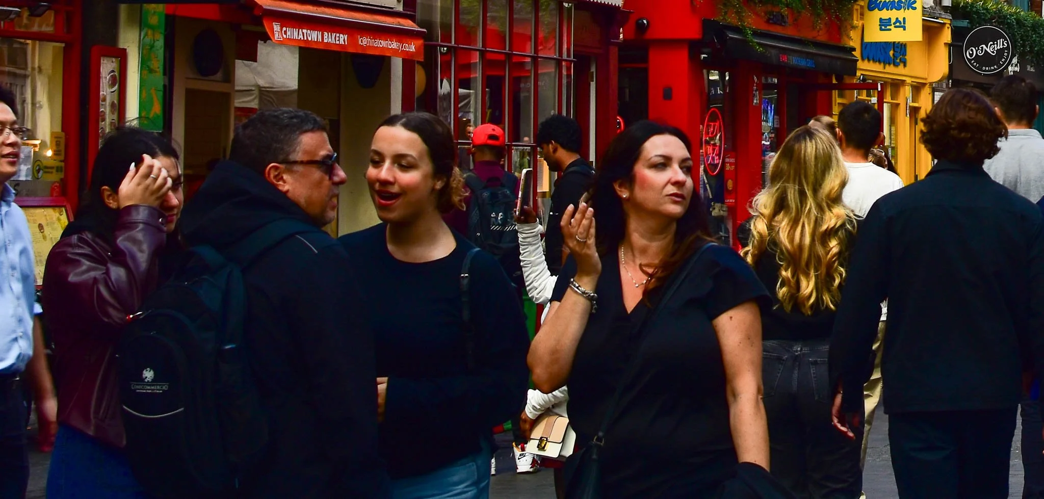Crowd of people walking on a busy city street with colorful storefronts, including eateries and shops, during daytime.