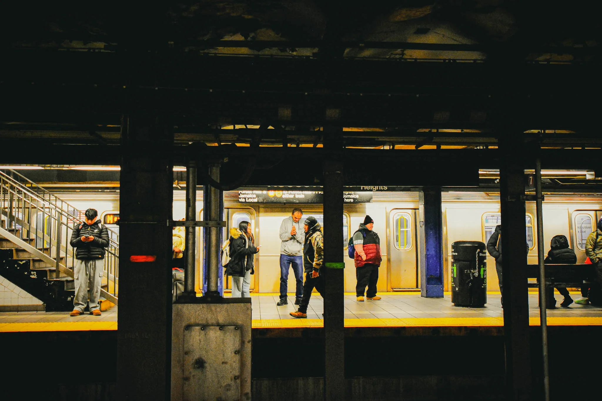 People waiting for a subway train at a station, with trains on both sides and some people standing, some looking at their phones, and others talking.