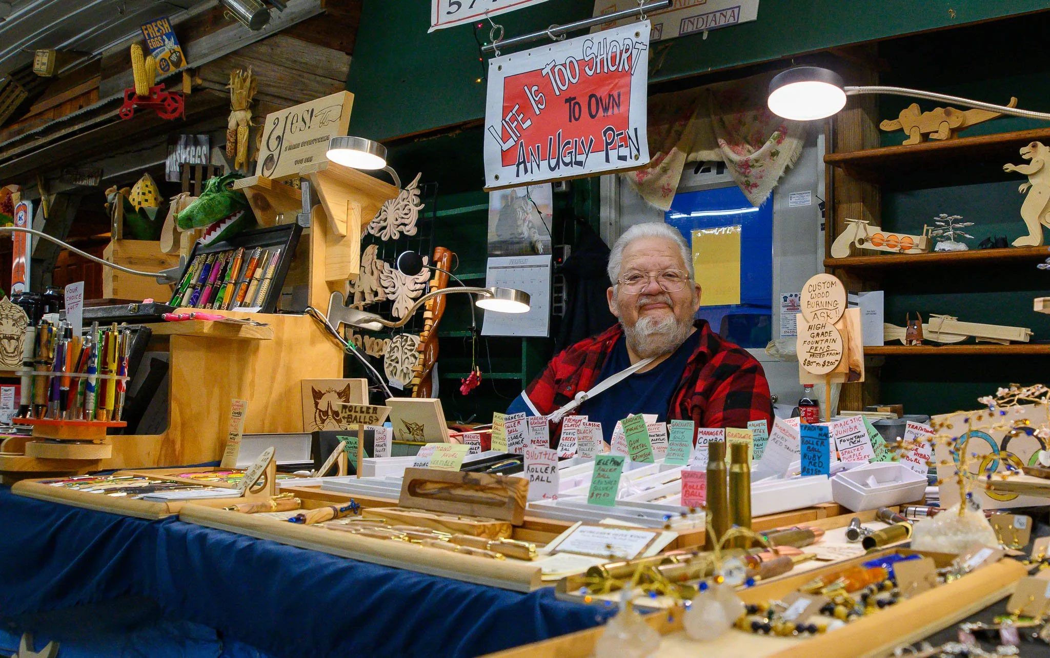 An elderly man with white hair and a beard, wearing glasses and a red plaid shirt, sitting at a booth with wooden crafts and jewelry for sale. Behind him is a sign that reads "Life is too short to own an ugly pen." The booth displays various wooden i