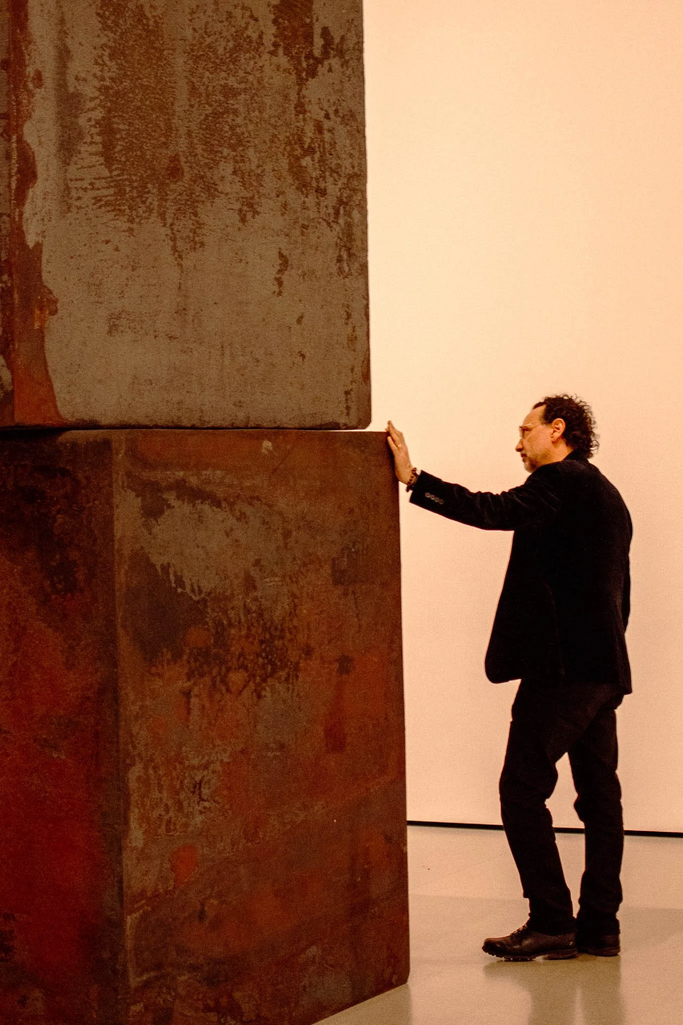 A man in a black suit touches a large rusted metal sculpture in an art gallery.