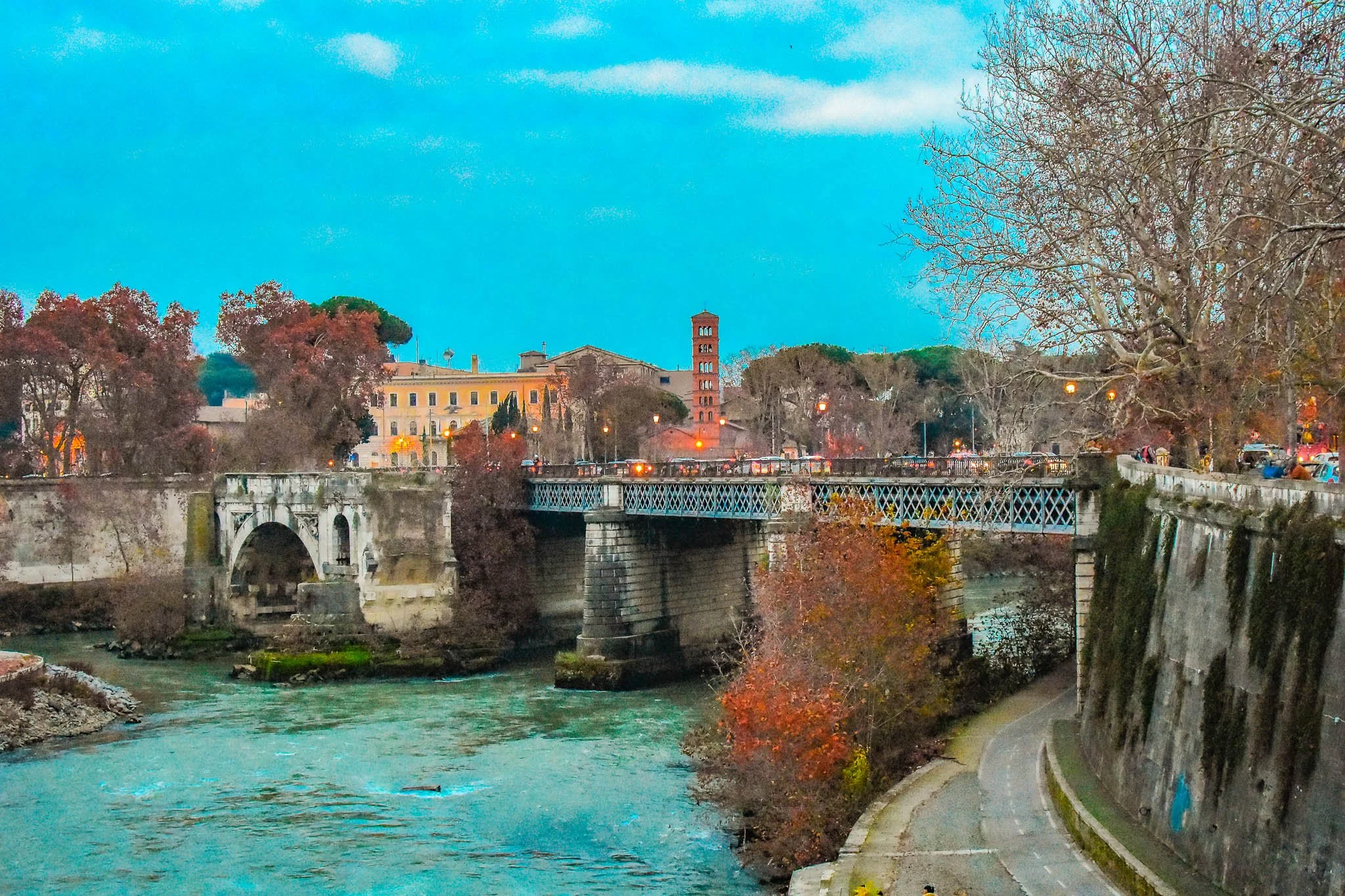A river flowing through a city with a bridge crossing over it, surrounded by trees with fall foliage and historic buildings in the background, under a partly cloudy sky.