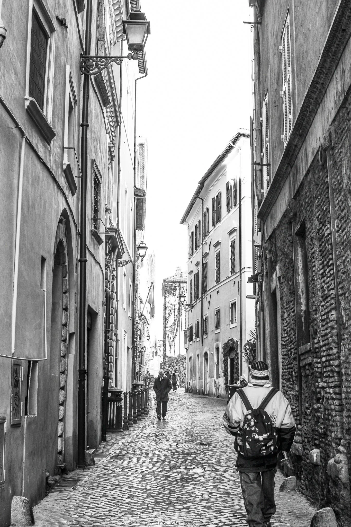 A narrow cobblestone street in a European city with old buildings on both sides. Two pedestrians, one with a backpack and another walking ahead, are visible. The street is lined with street lamps and windows with shutters.
