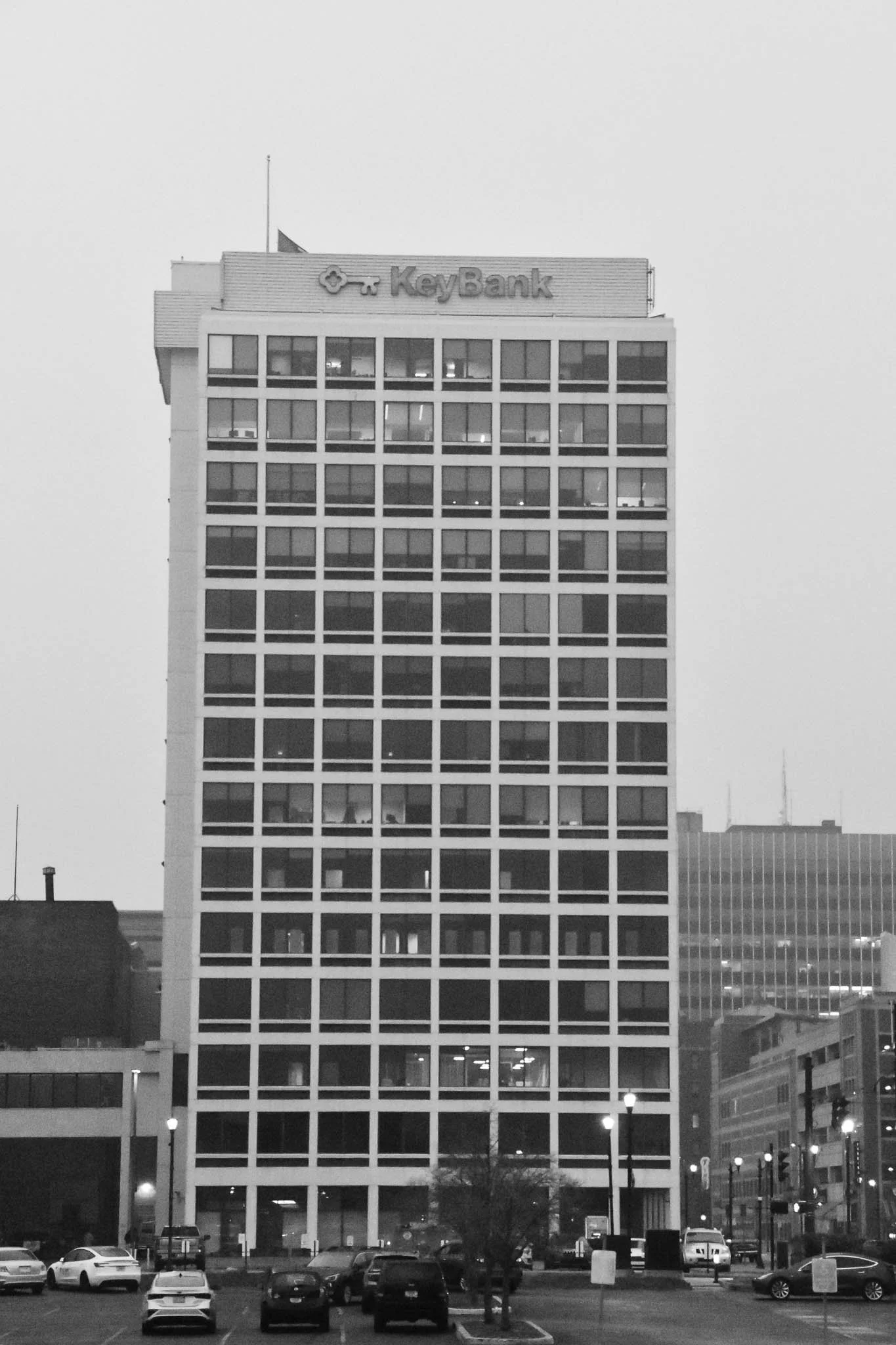 A tall office building with the KeyBank logo at the top. The building has a grid of windows, some with lights on. The foreground shows a parking lot with parked cars and a few street lamps.