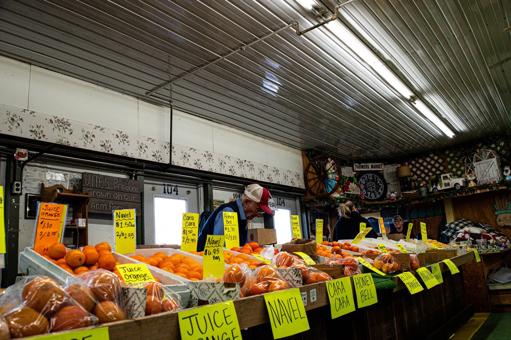 Indoor farmers market with various oranges and citrus fruits on display and yellow price tags. Customers browsing and a man wearing a blue jacket and white baseball cap shopping.
