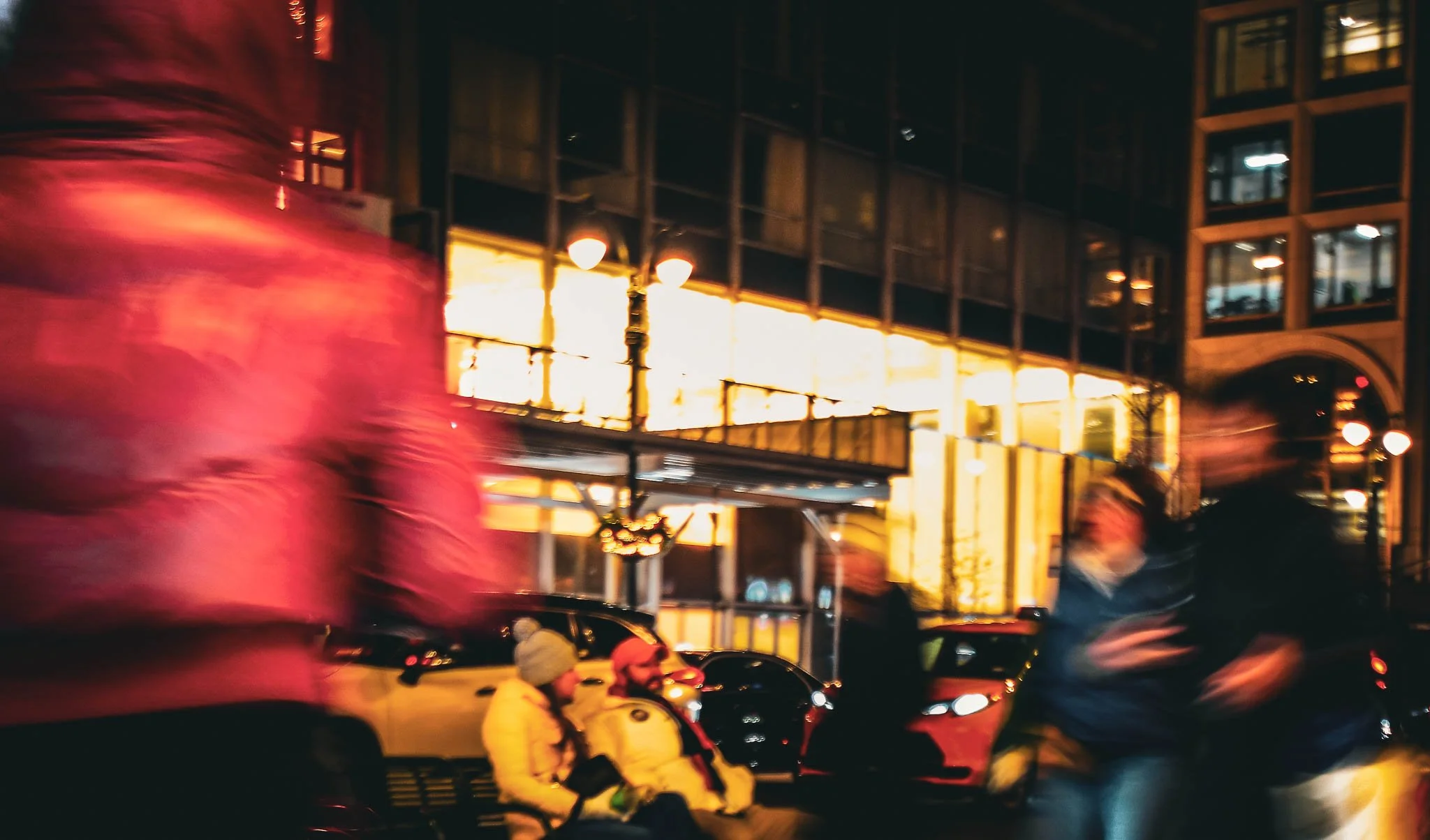 Blurred pedestrians walking on a city street at night, with a brightly lit building and parked cars in the background.