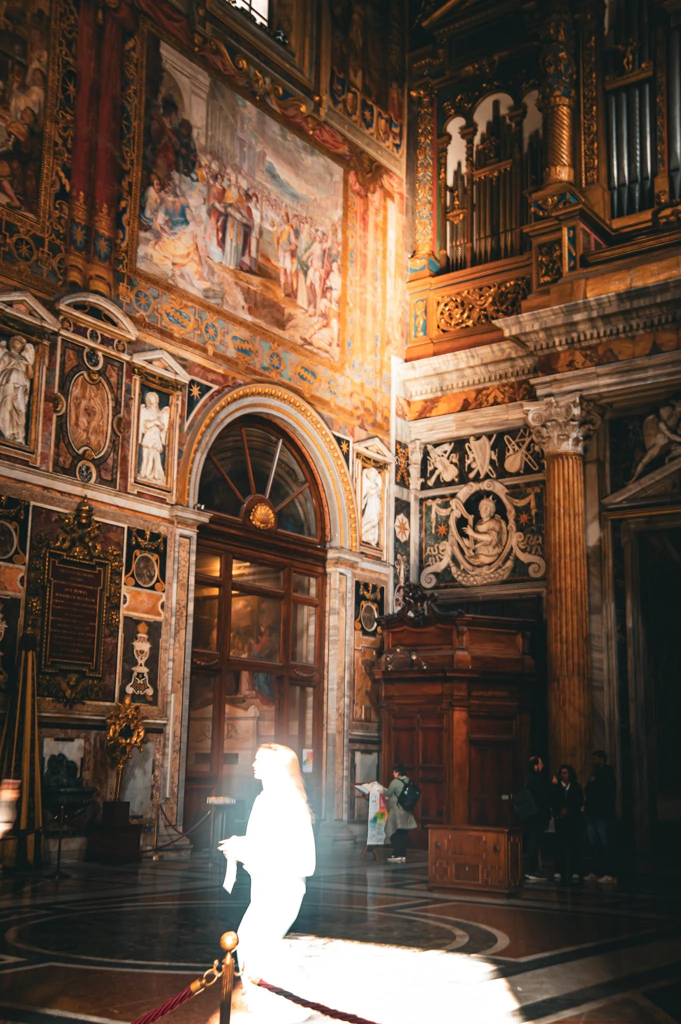 Interior of a richly decorated church or cathedral with ornate walls, frescoes, and woodwork, illuminated by natural light streaming through openings. Several people are inside, some reading or standing.
