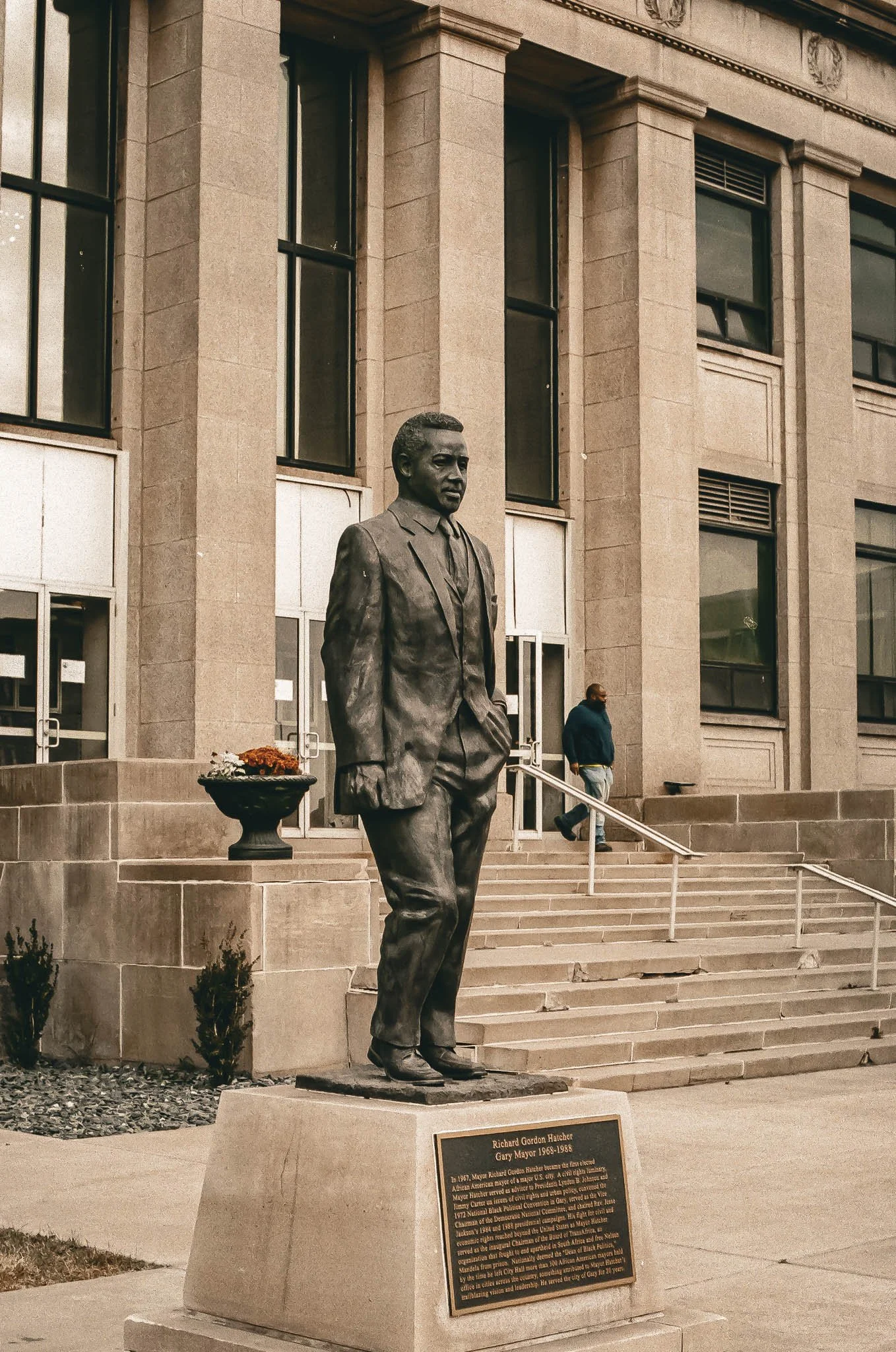 Bronze statue of a man in a suit, standing outside a government building. The statue is placed on a concrete pedestal with a plaque. The background shows a stone building with large windows and steps leading up to the entrance.