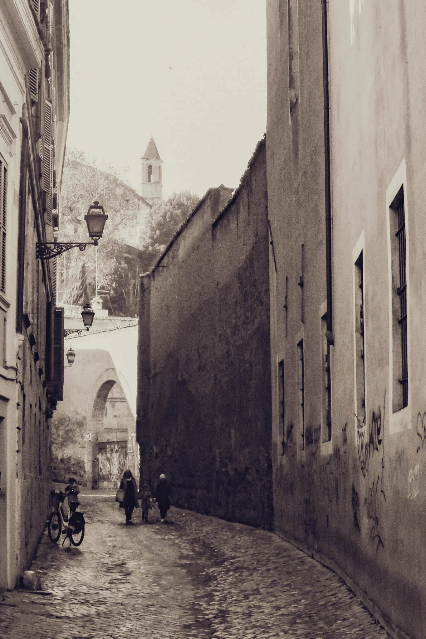 A black and white photo of a narrow cobblestone street in an old European city. There are three people walking away from the camera, two adults and a child. On the left, there are old buildings with shutters and street lamps. There is a bicycle leani