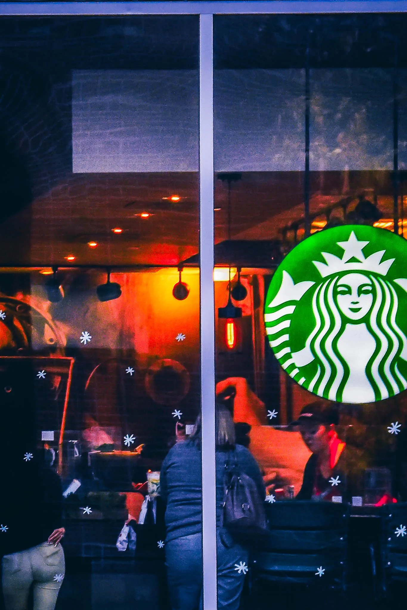 View of a Starbucks cafe window with a green Starbucks logo, reflective interior lighting, and several people inside sitting and standing.