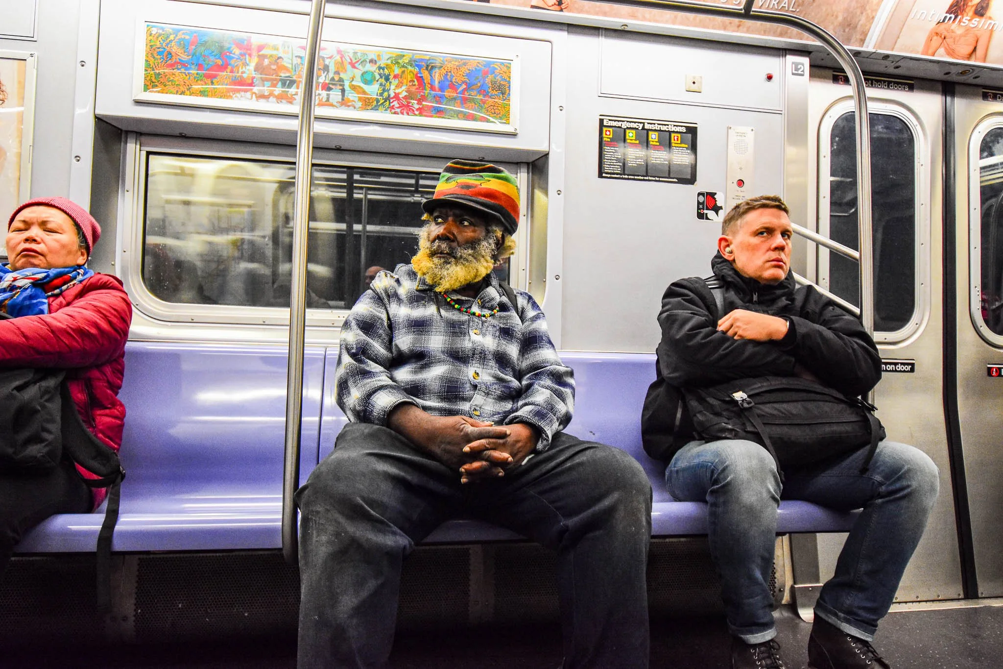 Three passengers seated on a subway train, including a man with a colorfully patterned hat, a woman with closed eyes wearing a red jacket, and a young man with a serious expression wearing a black jacket and jeans.