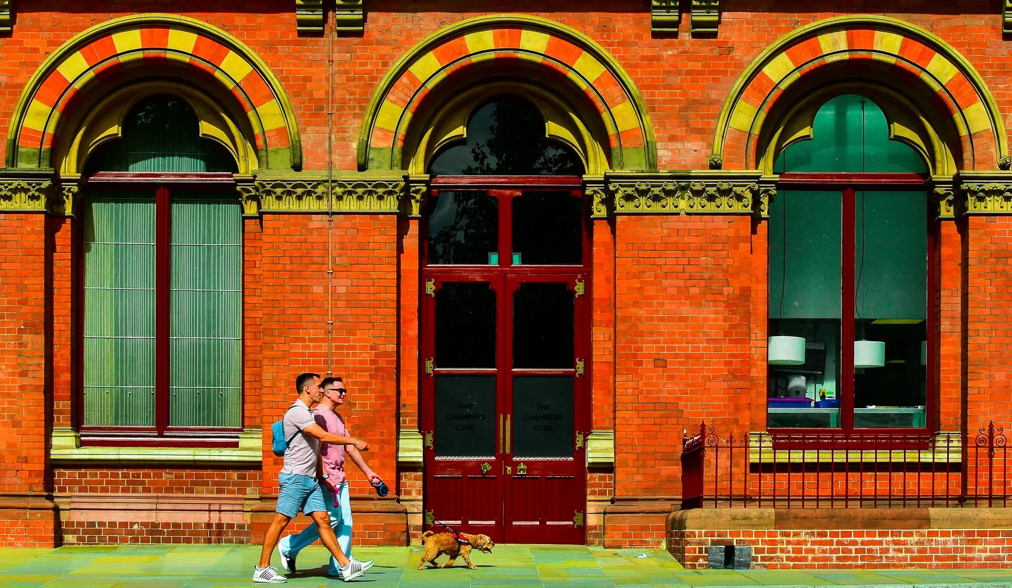 Two men walking a dog outside a red brick building with large arched windows. The building has decorative yellow and red trim around the windows and entrance doors with the sign 'The Chambers Club'.