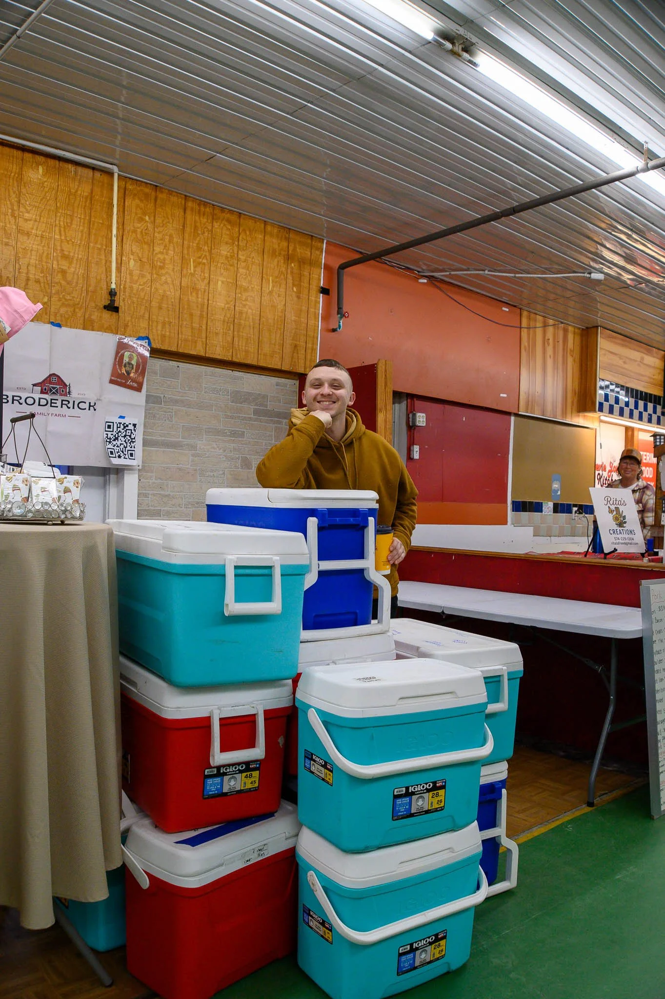 A smiling man in a mustard-colored hoodie standing behind stacked coolers inside a building with wooden and brick walls. A woman in the background is at a booth with a sign that reads 'Ritas Creations'.