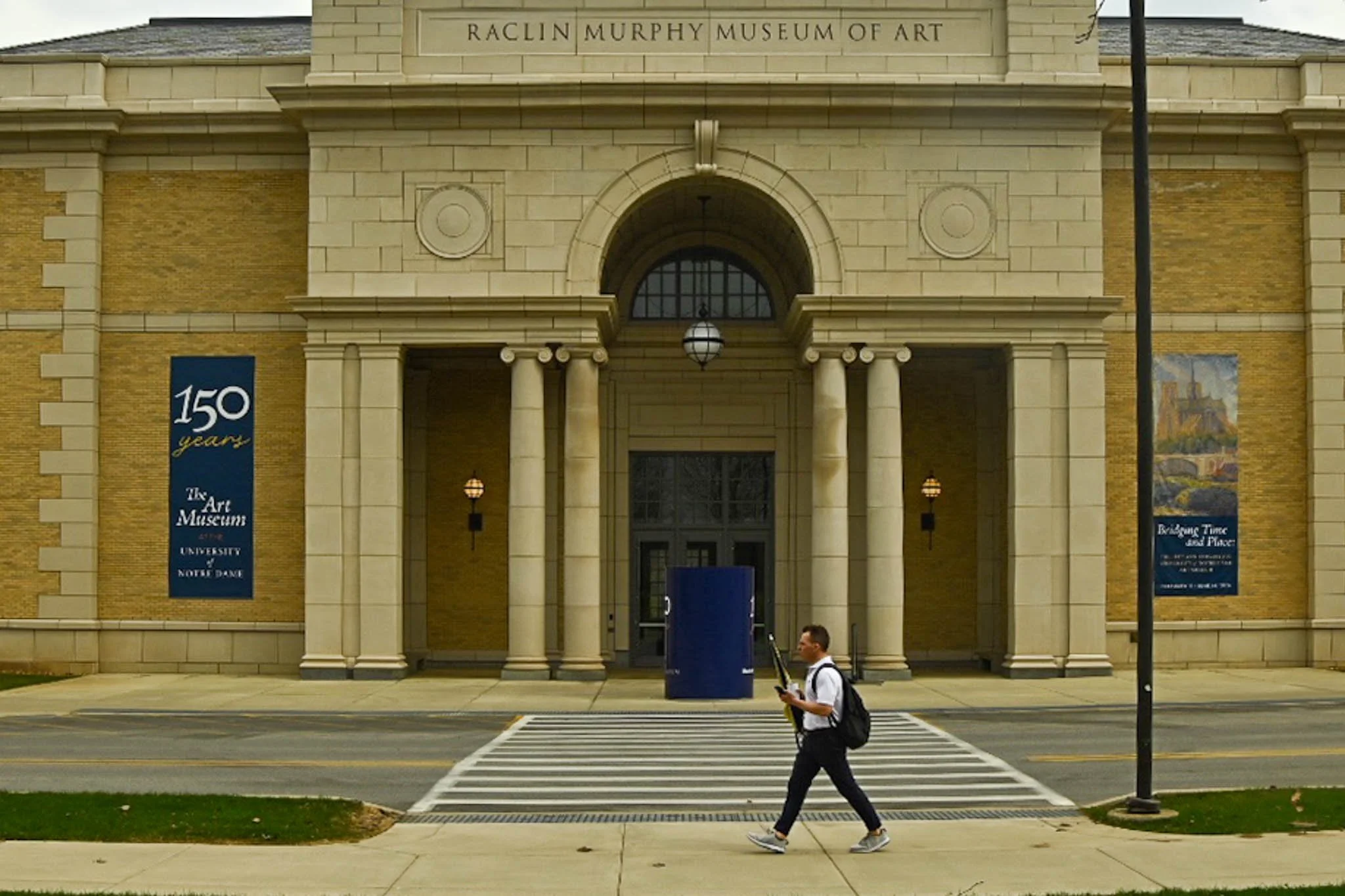 The facade of the Raclin Murphy Museum of Art at the University of Notre Dame with a man walking across the crosswalk in front.