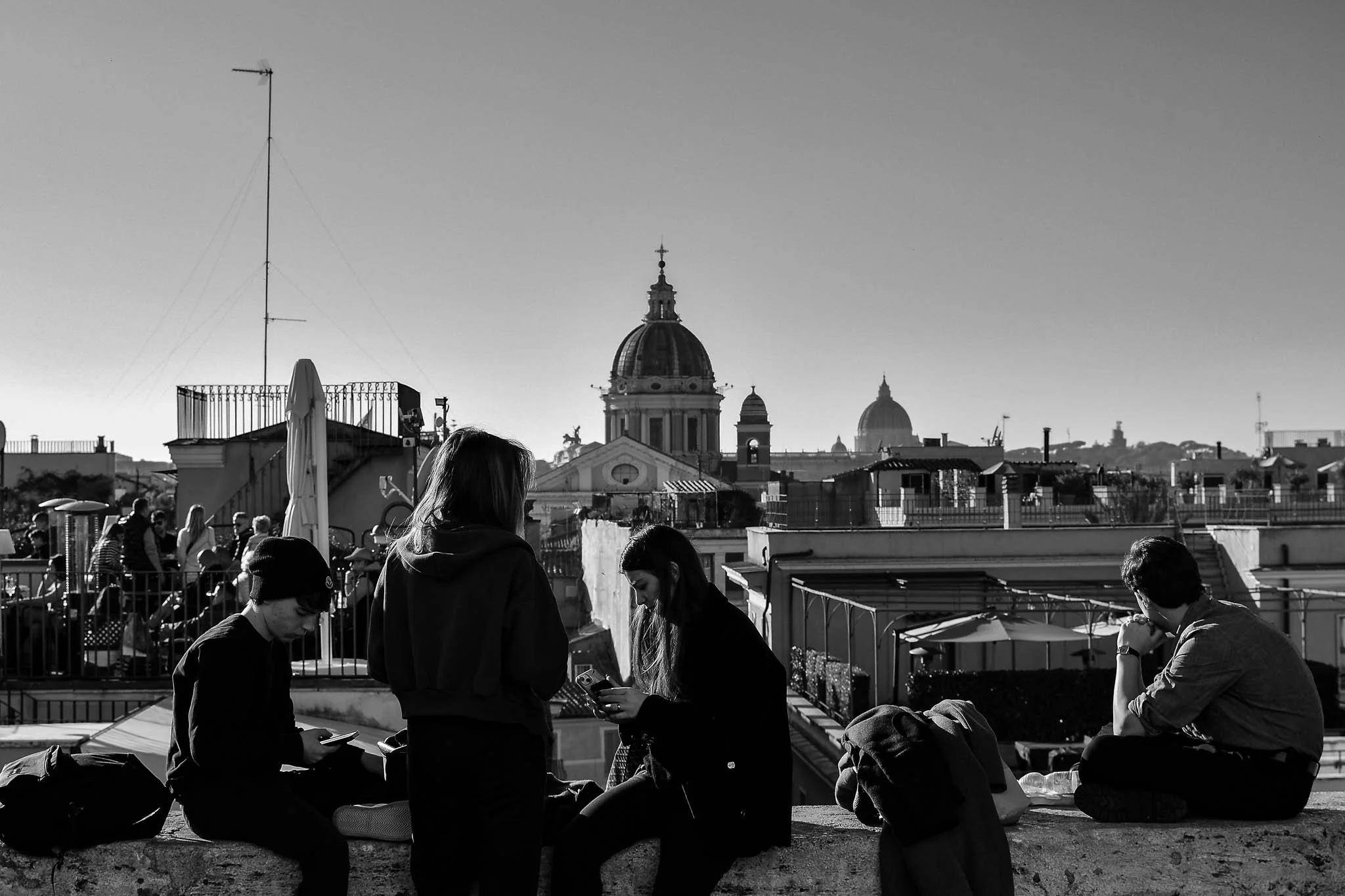 Black and white photo of four young people on a rooftop in a city with iconic domed and spired buildings in the background, some sitting and some standing, all using their phones.