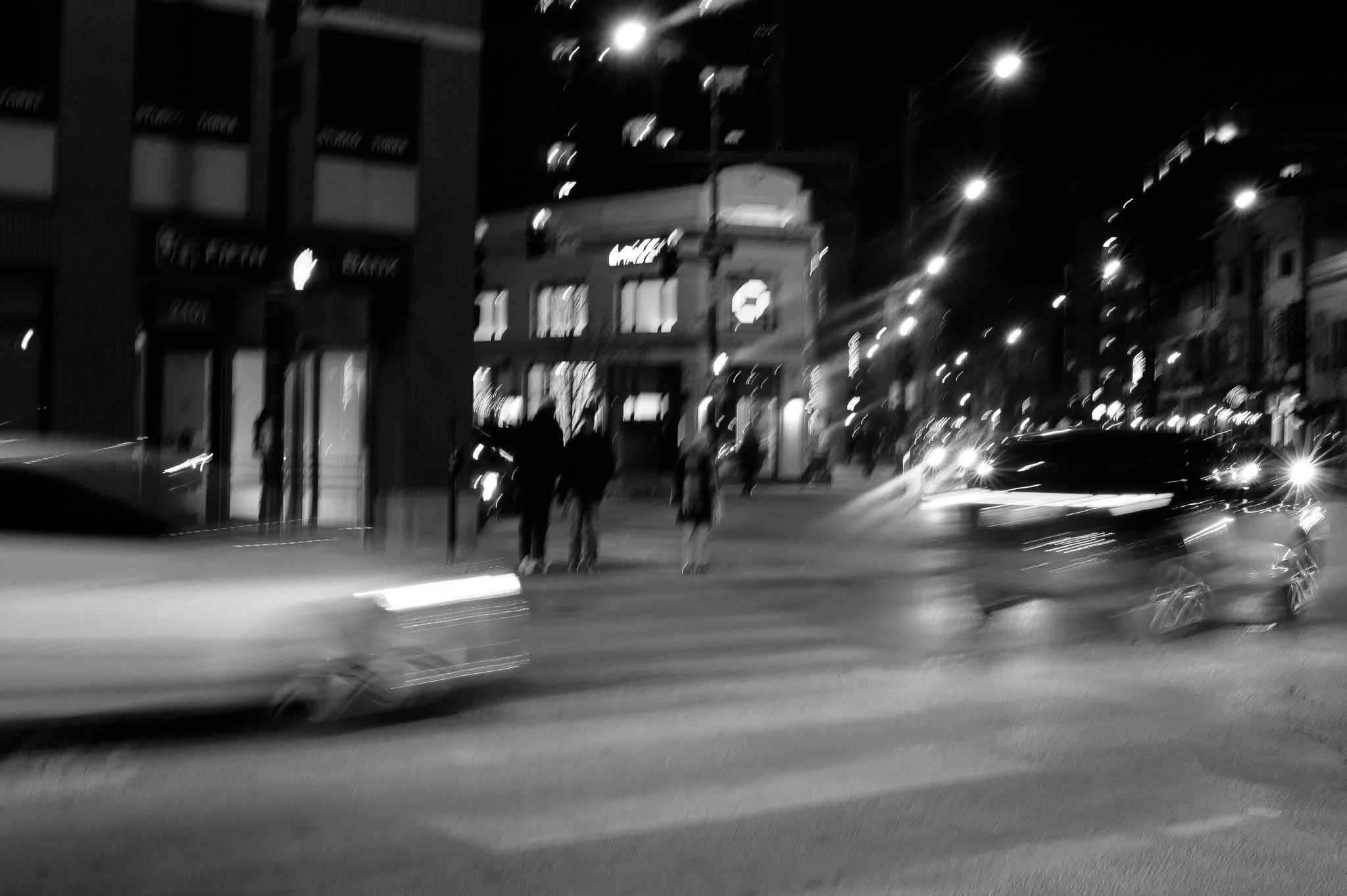 Nighttime city street scene in black and white with people crossing the street, blurred cars, building storefronts, and streetlights.