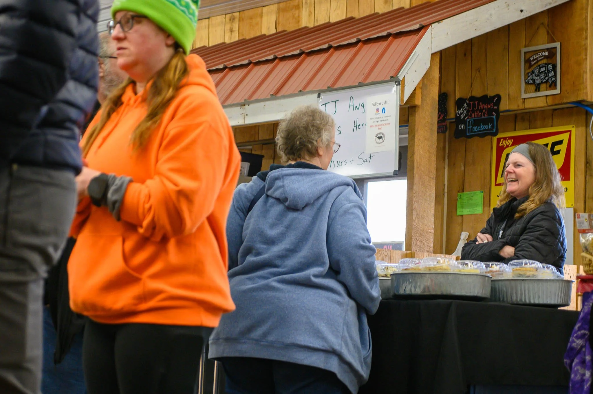 Three women are at a food counter inside a wooden building. One woman, in an orange hoodie and green knit cap, is standing in front, while two women are behind the counter. One of them, with glasses and curly hair, is in a blue hoodie, and the other,