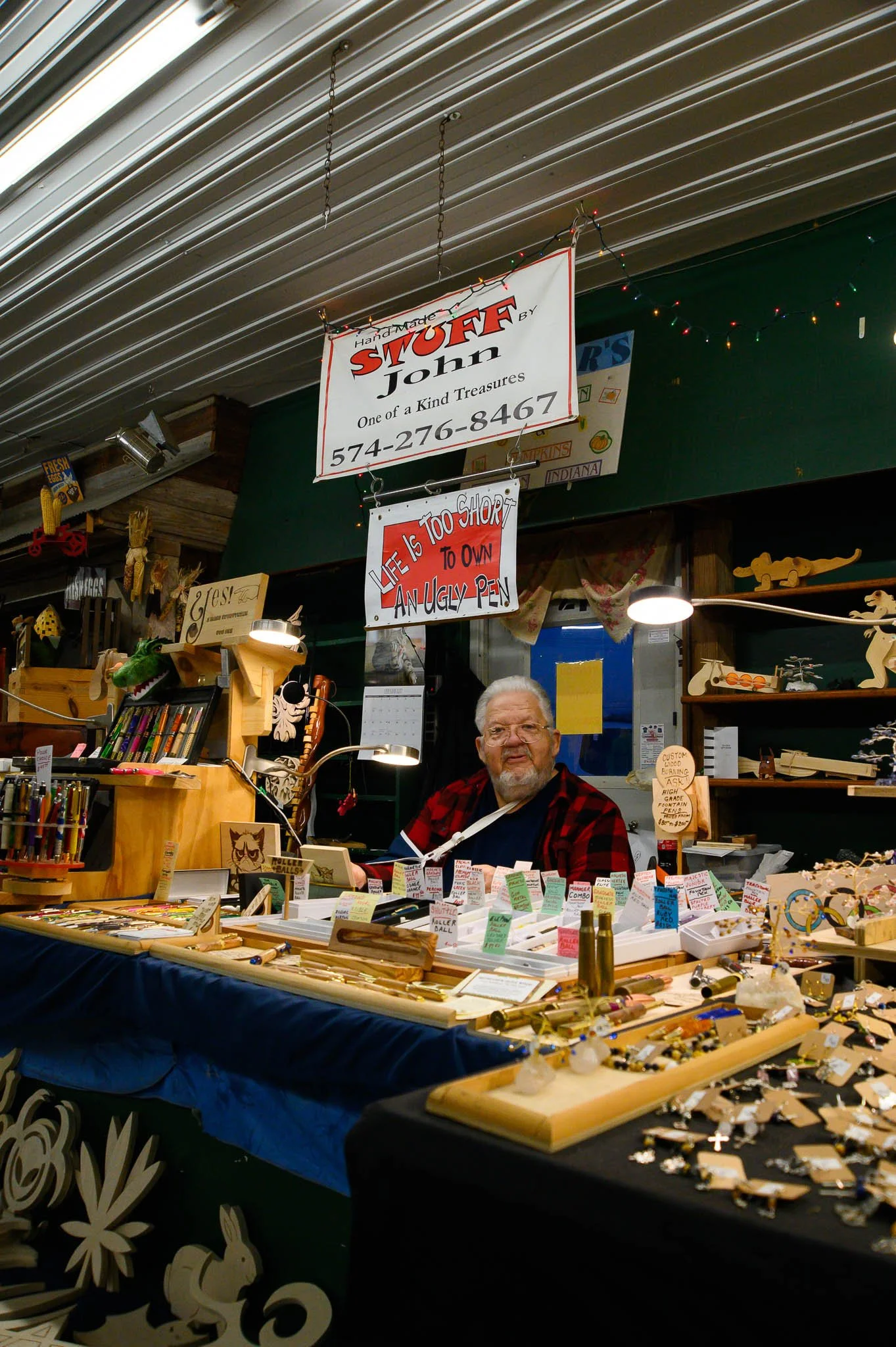 A man with glasses and a beard sitting at a craft booth filled with handmade jewelry, wooden crafts, and signs, under a ceiling with string lights.