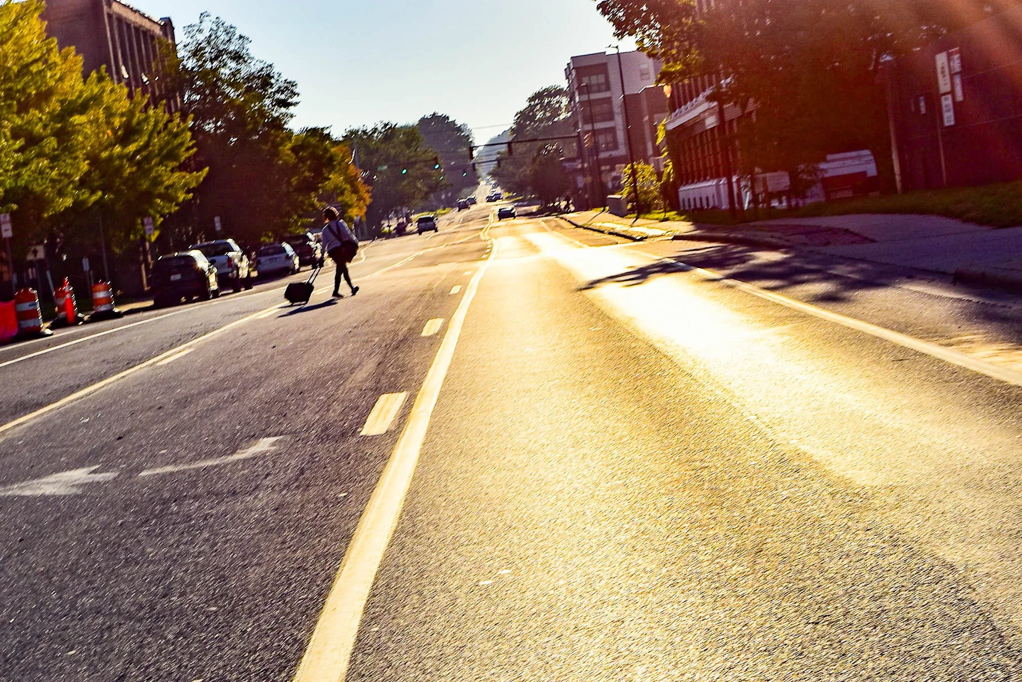 A city street during sunset, with a person walking and pulling a suitcase in the middle of the street. Parked cars line the left side, and apartment buildings and trees are on the right. Traffic lights are visible in the distance.